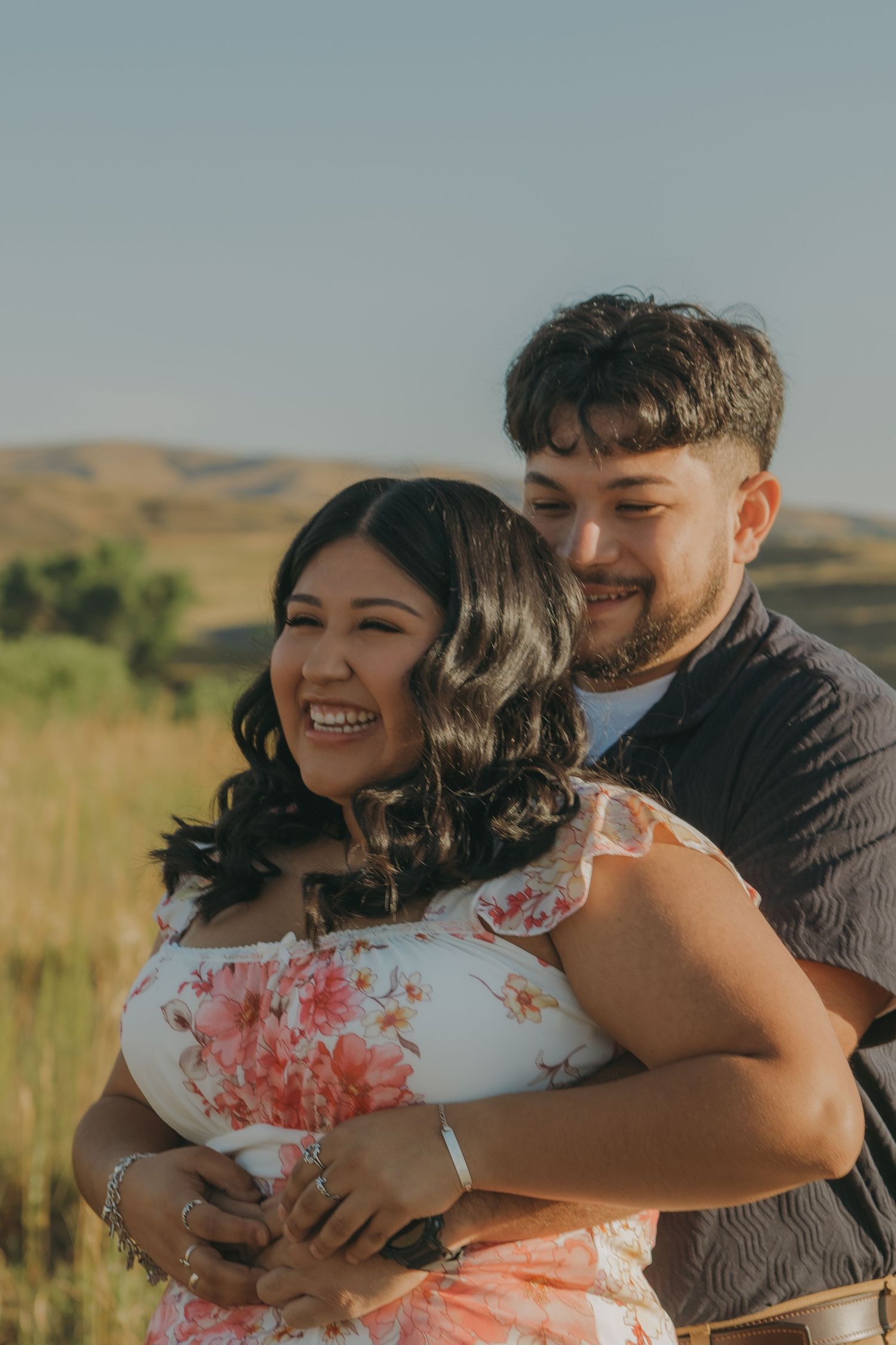 A couple embraces in a warm golden field with rolling hills in the background.