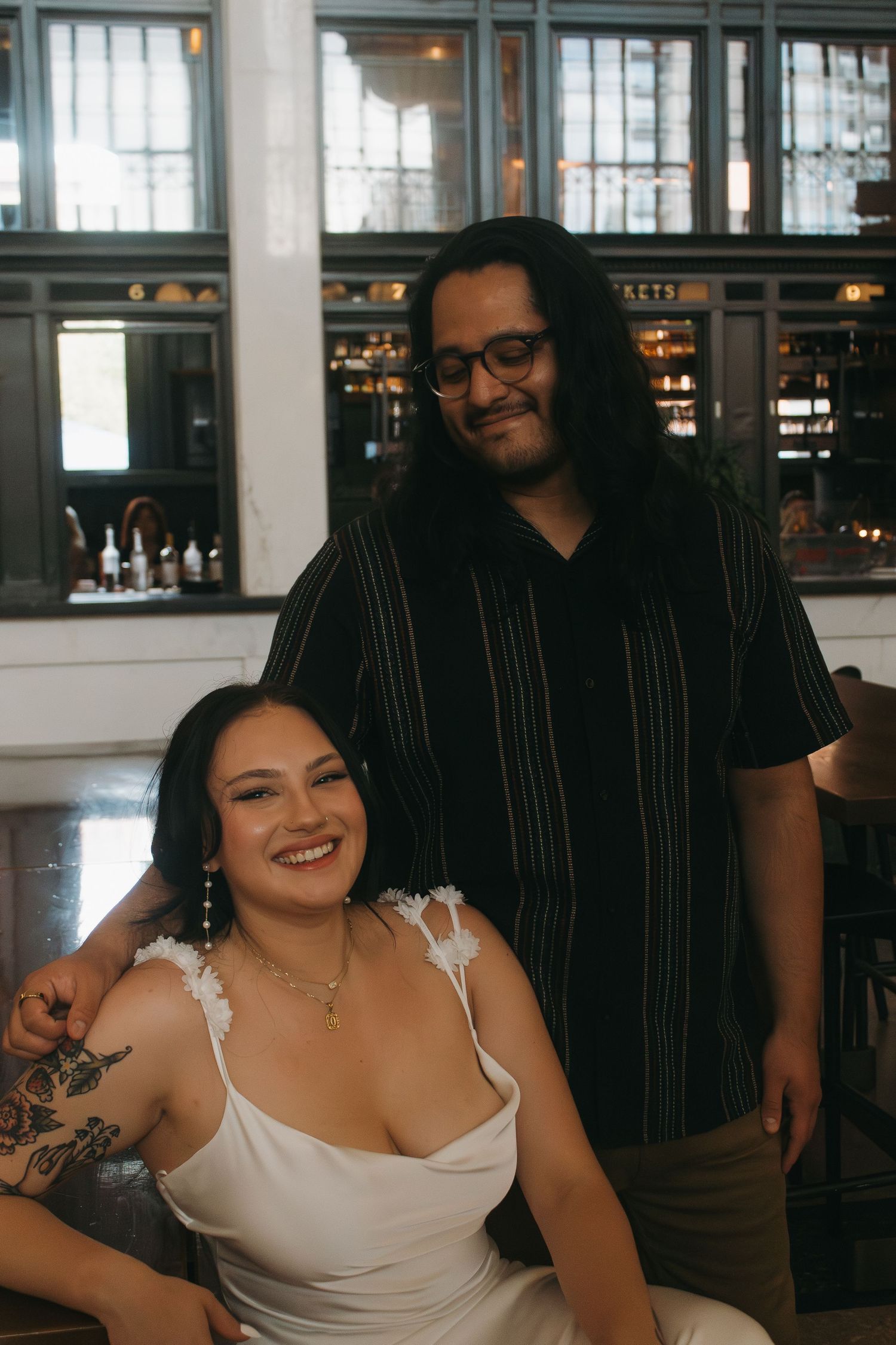 Two people posing together in a restaurant, one wearing a striped shirt and one in a white dress with tattoos.