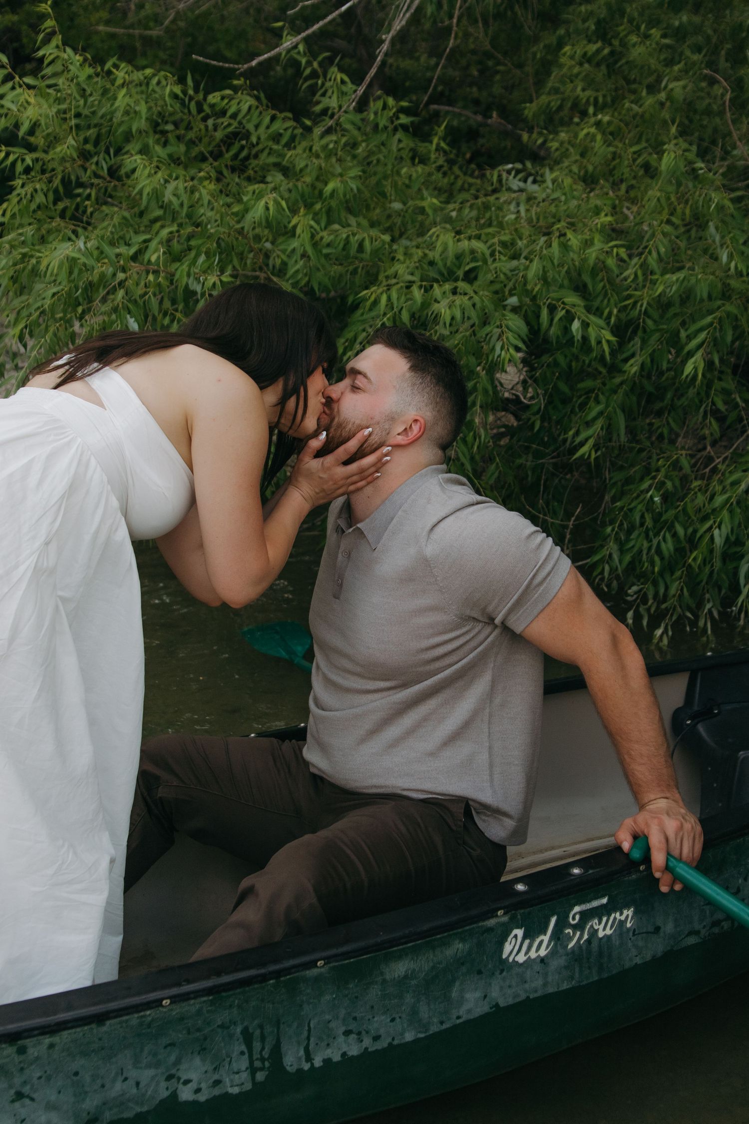 A couple shares an intimate moment aboard a dark canoe surrounded by overhanging branches.