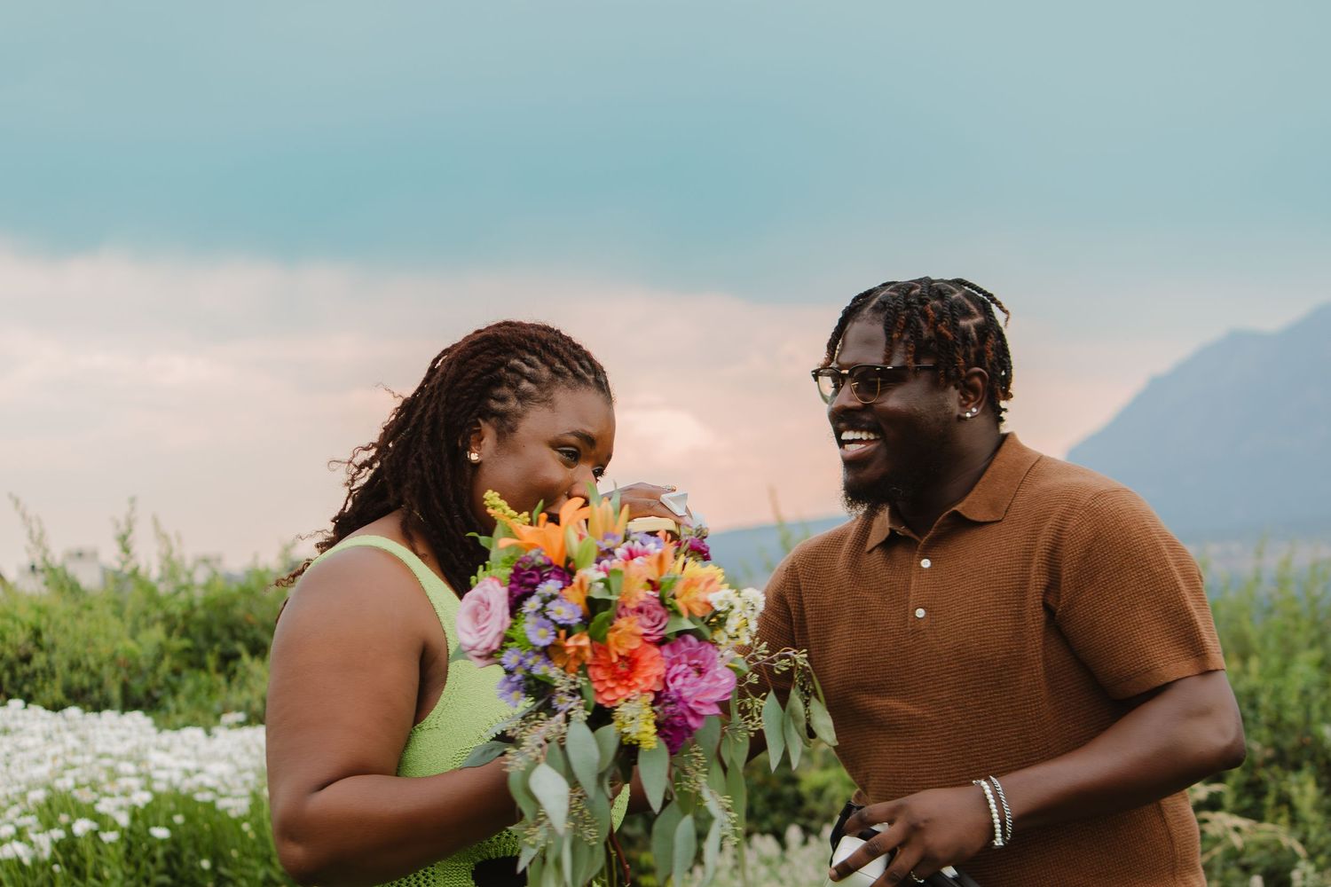 A couple shares a joyful moment with colorful wildflowers in a scenic mountain meadow at sunset.