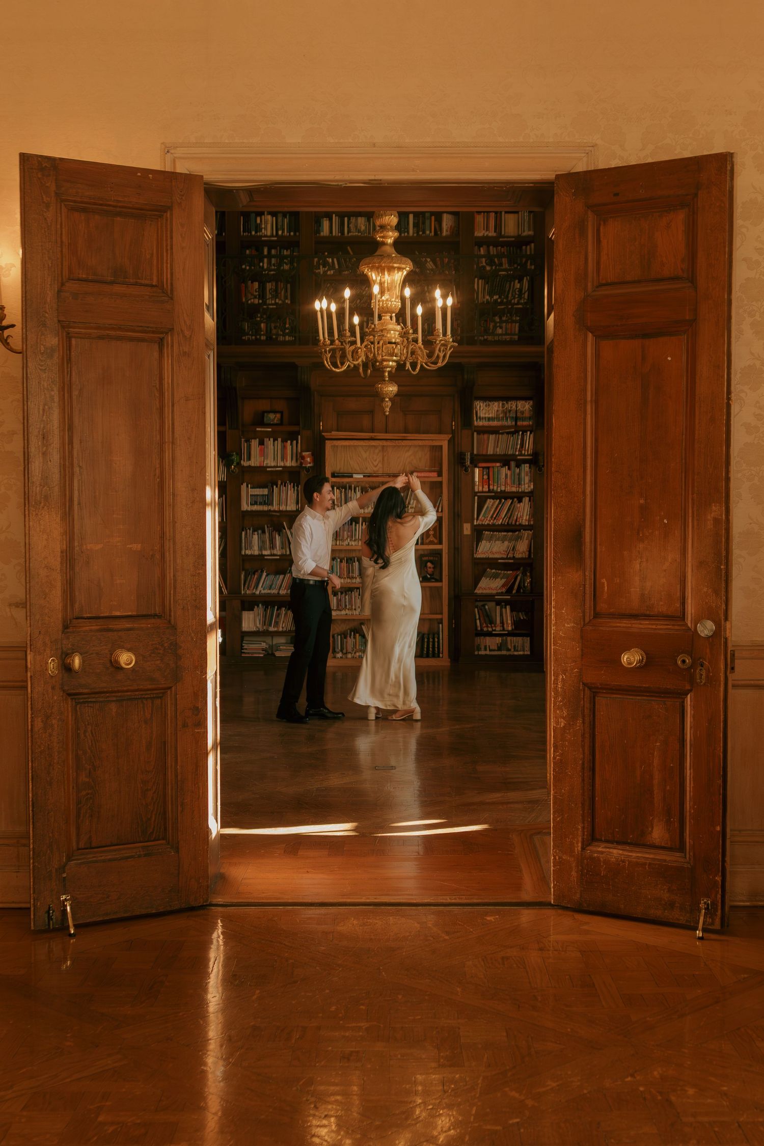 Two figures dance gracefully in an elegant wood-paneled library with bookshelves and a chandelier overhead.