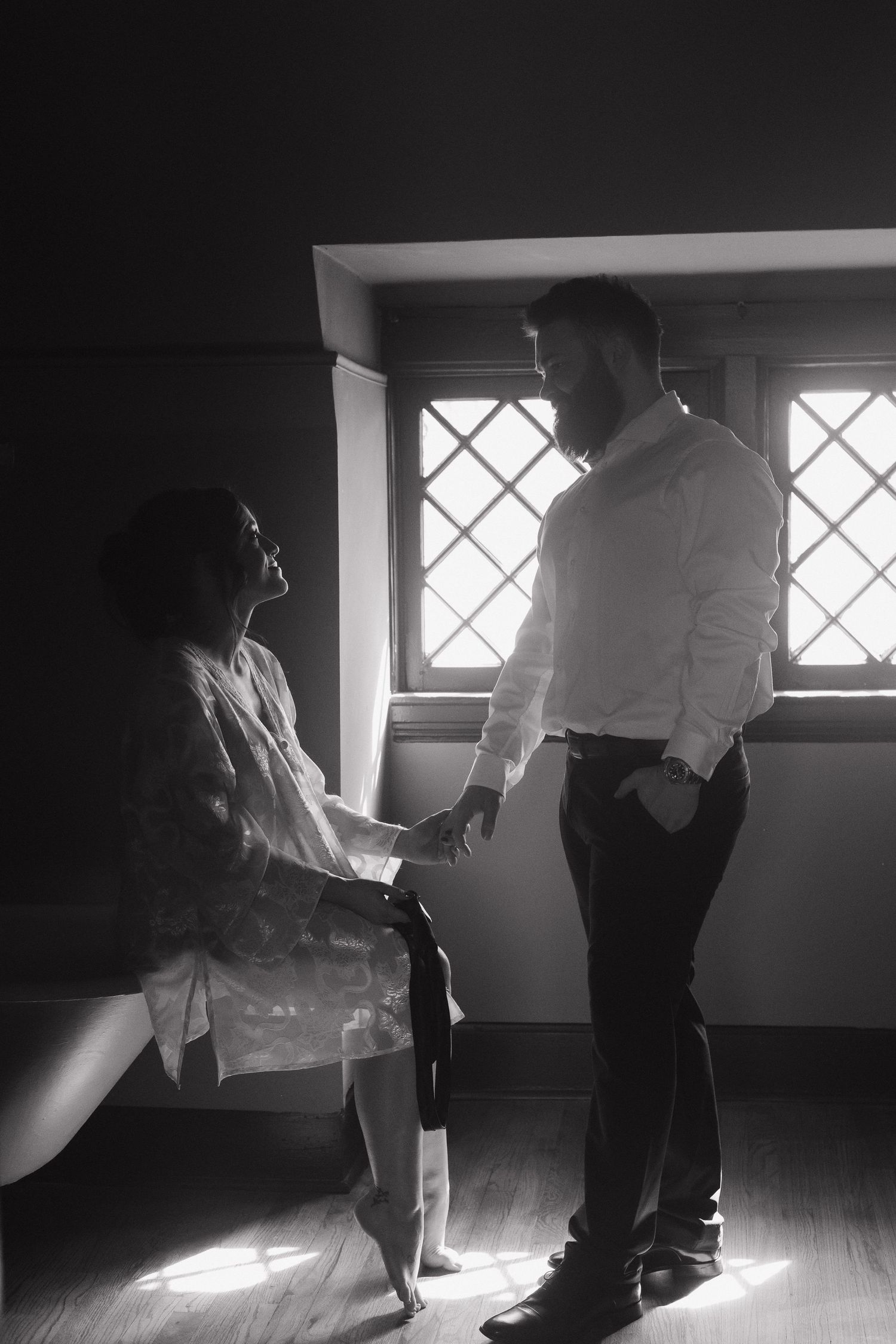 Moody black and white photo of couple sharing intimate moment by window with dramatic lighting.