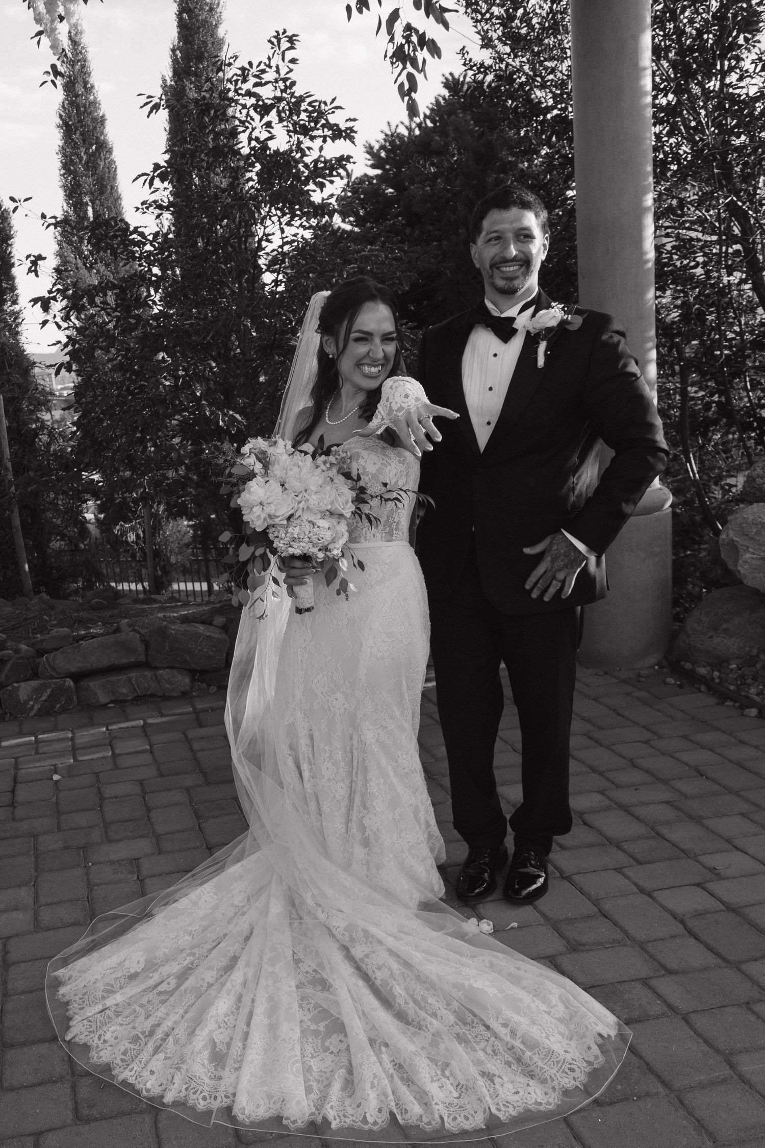 Joyful bride and groom pose together outdoors, bride holding bouquet in elegant lace gown and veil.