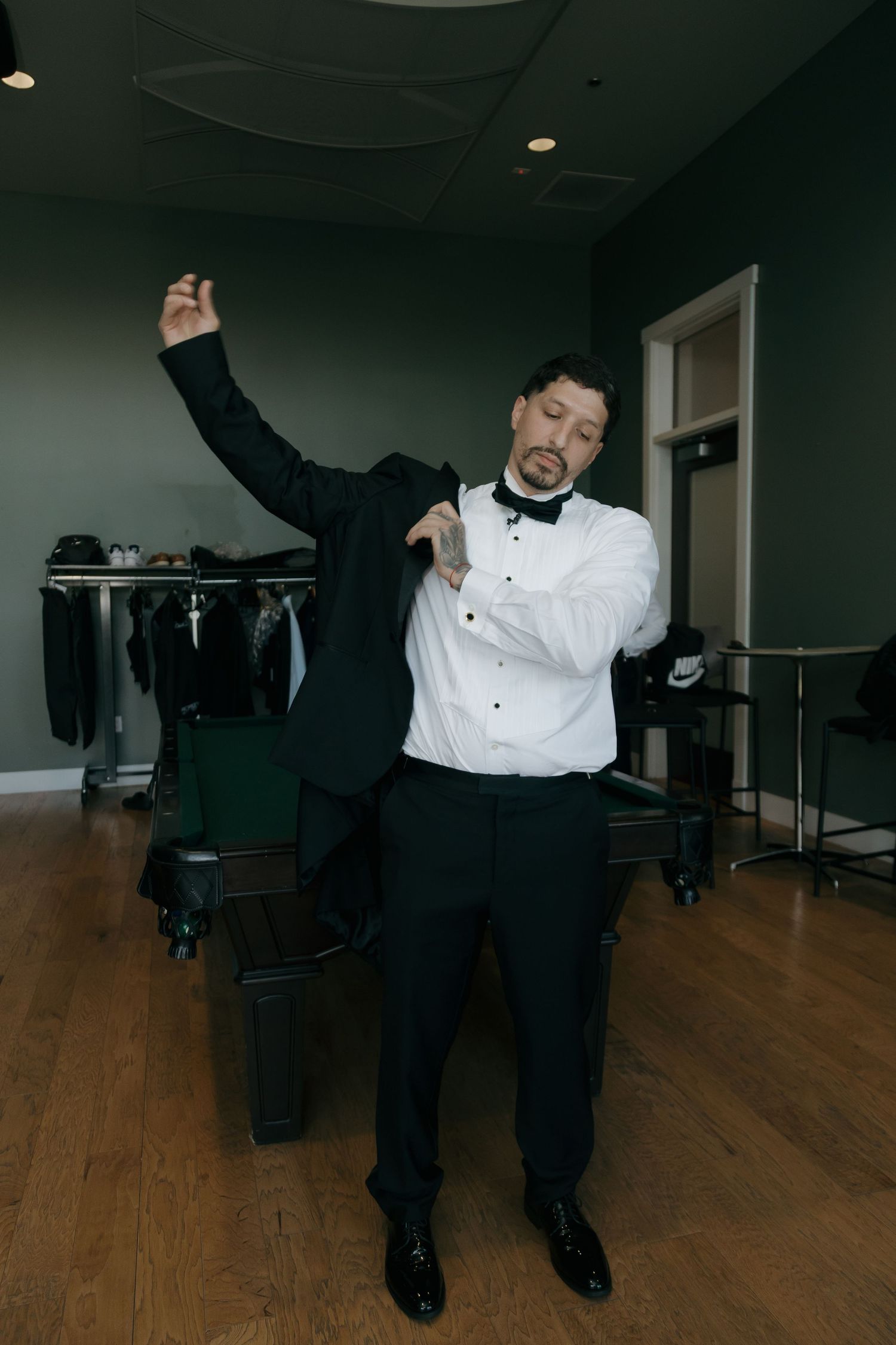 Groom in tuxedo playfully putting on his jacket in a green-walled wedding preparation room.