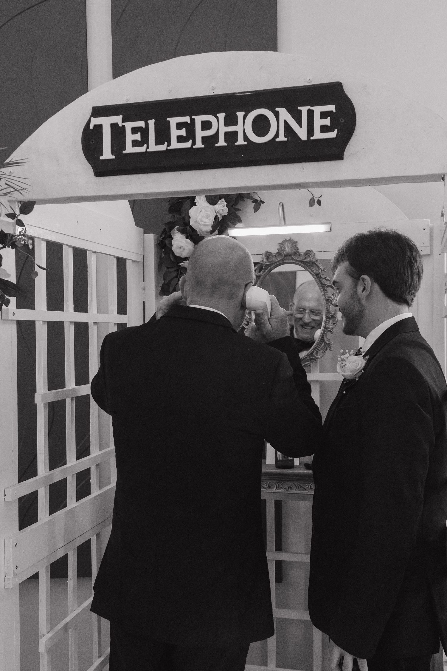 Black and white photo of groomsmen gathered at decorative telephone booth at wedding reception.