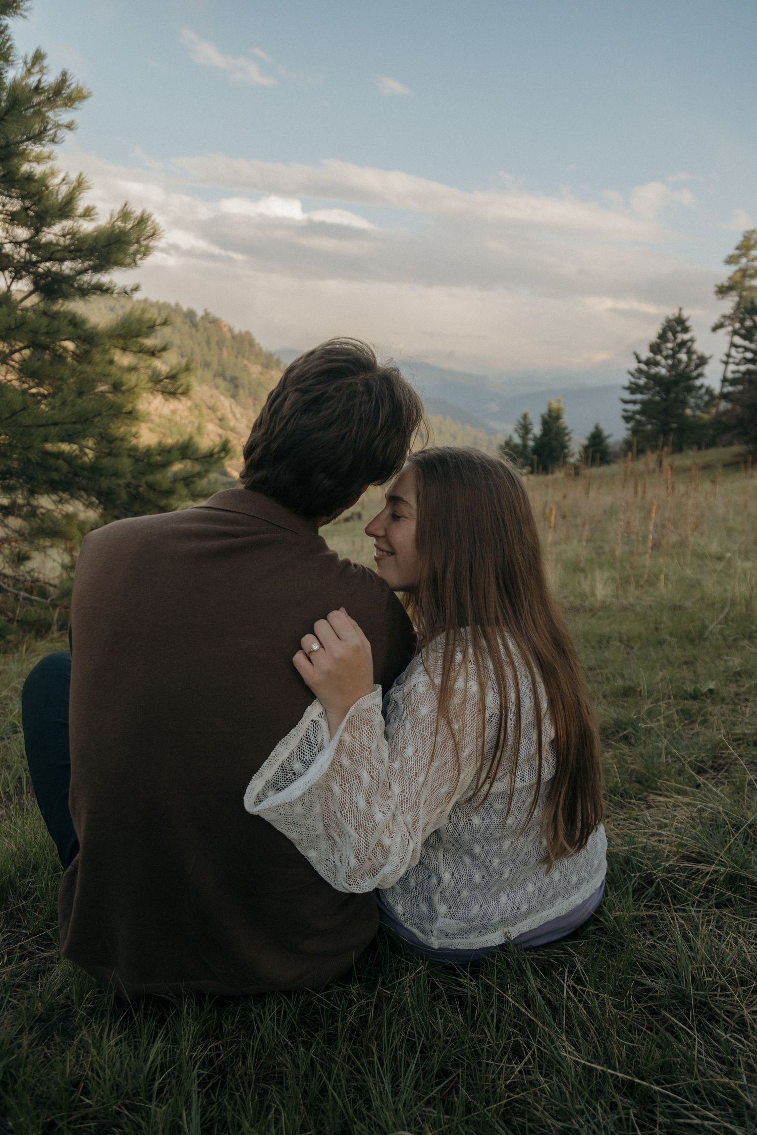 Romantic couple embracing while sitting on a grassy hillside with mountain views at dusk.