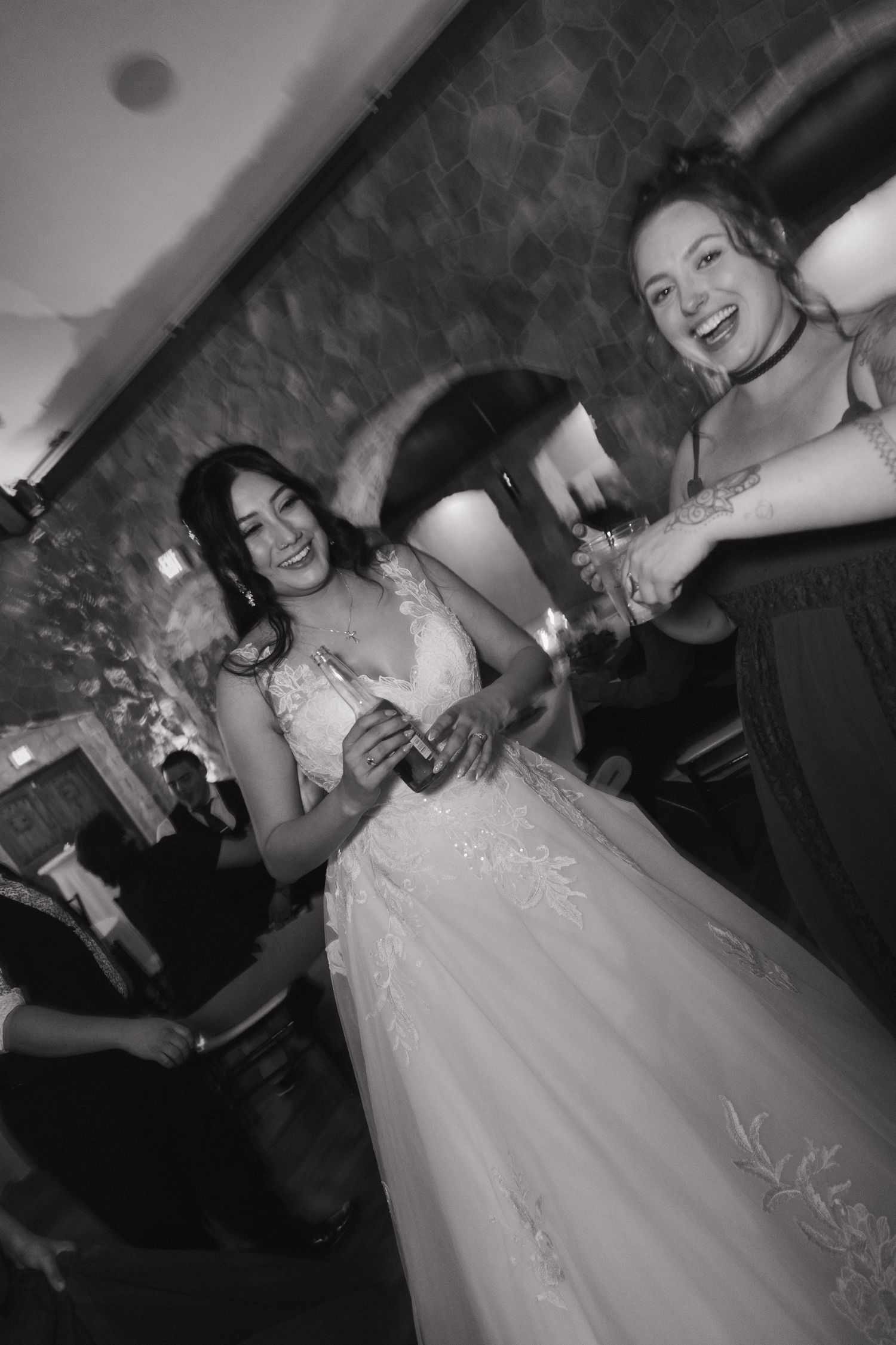 Black and white photo of bride in lace gown laughing with bridesmaid during wedding reception.