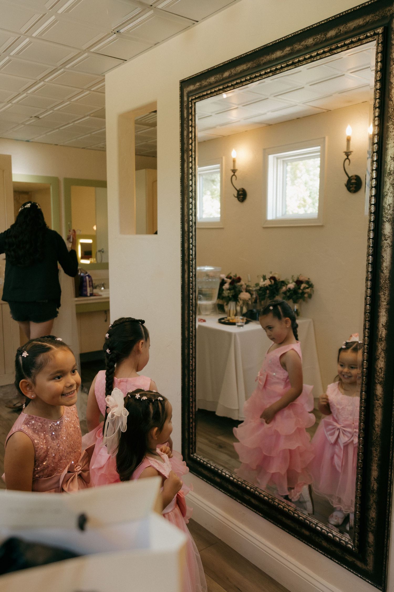 Young flower girls in pink dresses admiring their reflections in ornate mirror in bridal suite.