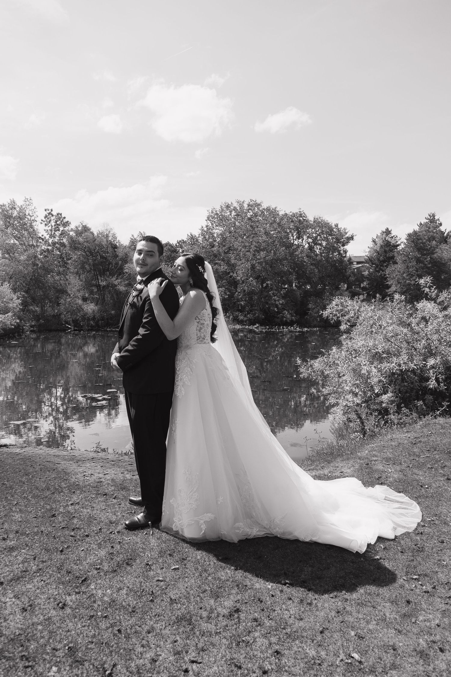 Black and white photo of bride and groom posing romantically by a serene lake with trees.