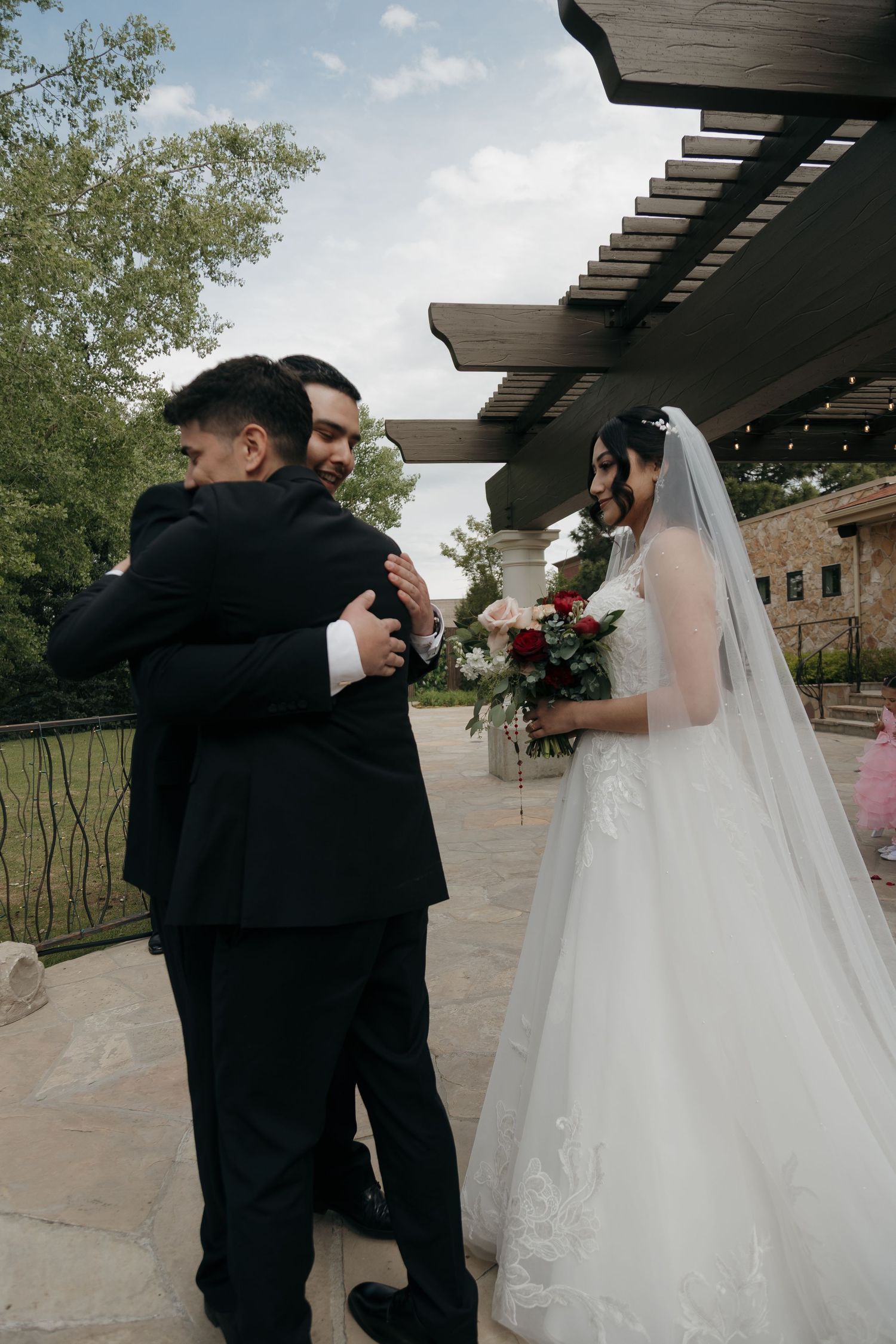 Groom embraces a guest while bride in white lace gown holds red rose bouquet nearby.