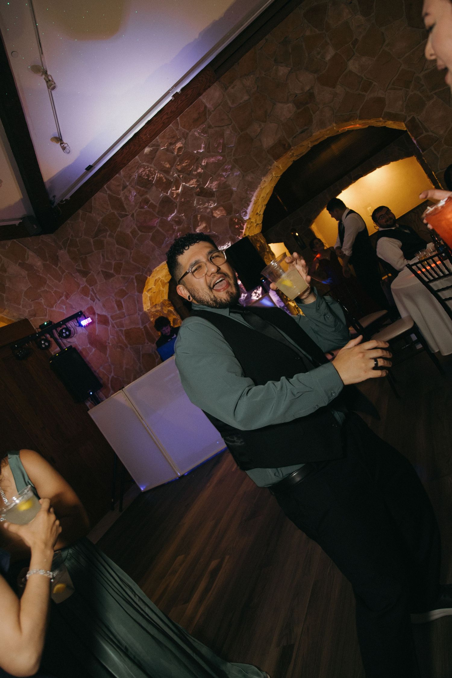 Man in green vest dances with a drink at a lively wedding reception with purple ambient lighting.
