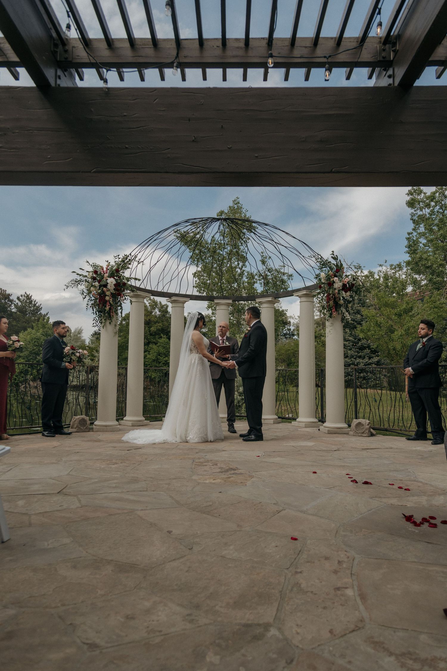 Bride and groom exchanging vows under a floral arch with white columns at an outdoor wedding ceremony.