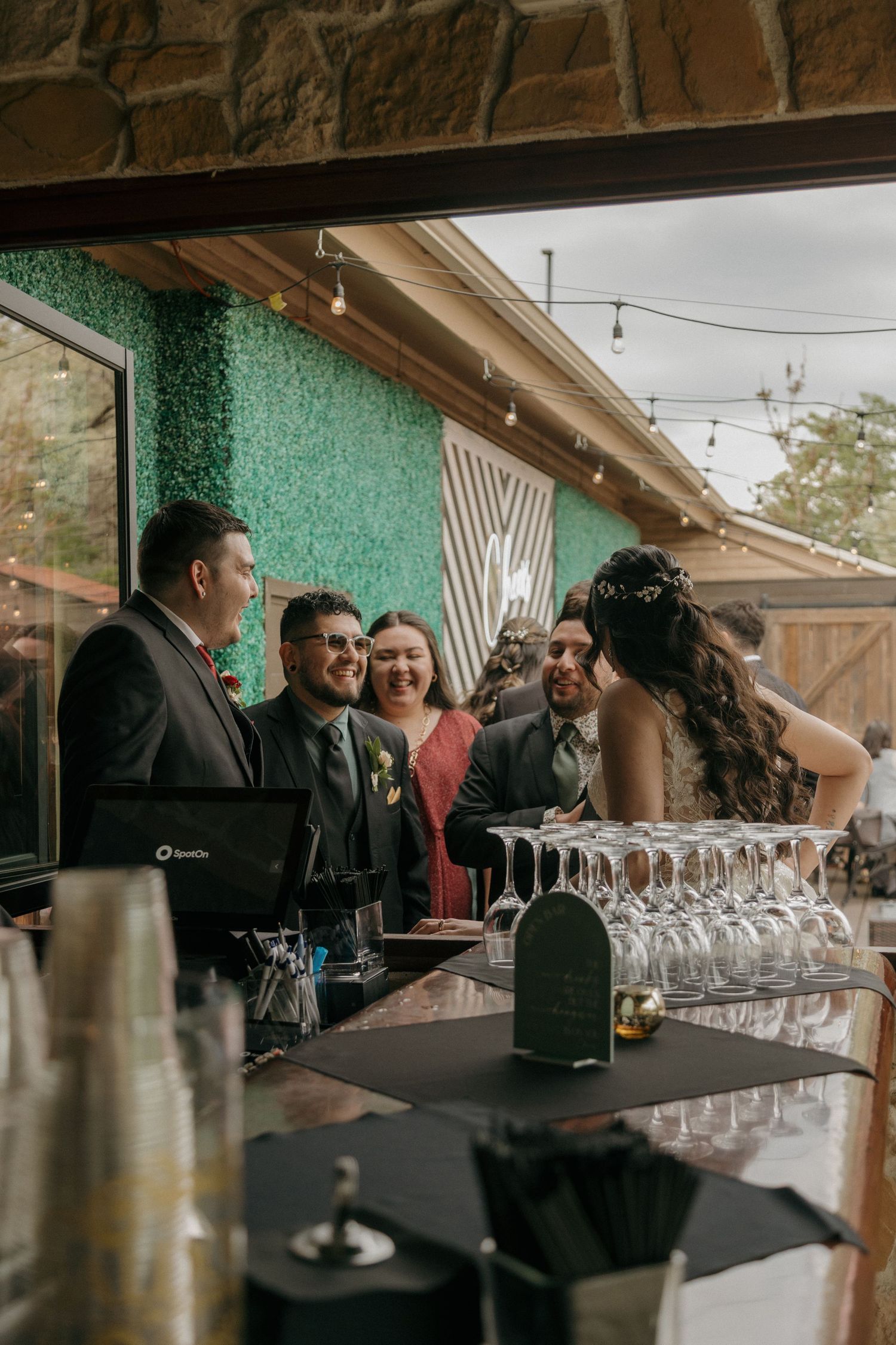 Wedding guests and the bridal couple laugh together at a bar adorned with a green hedge wall backdrop.