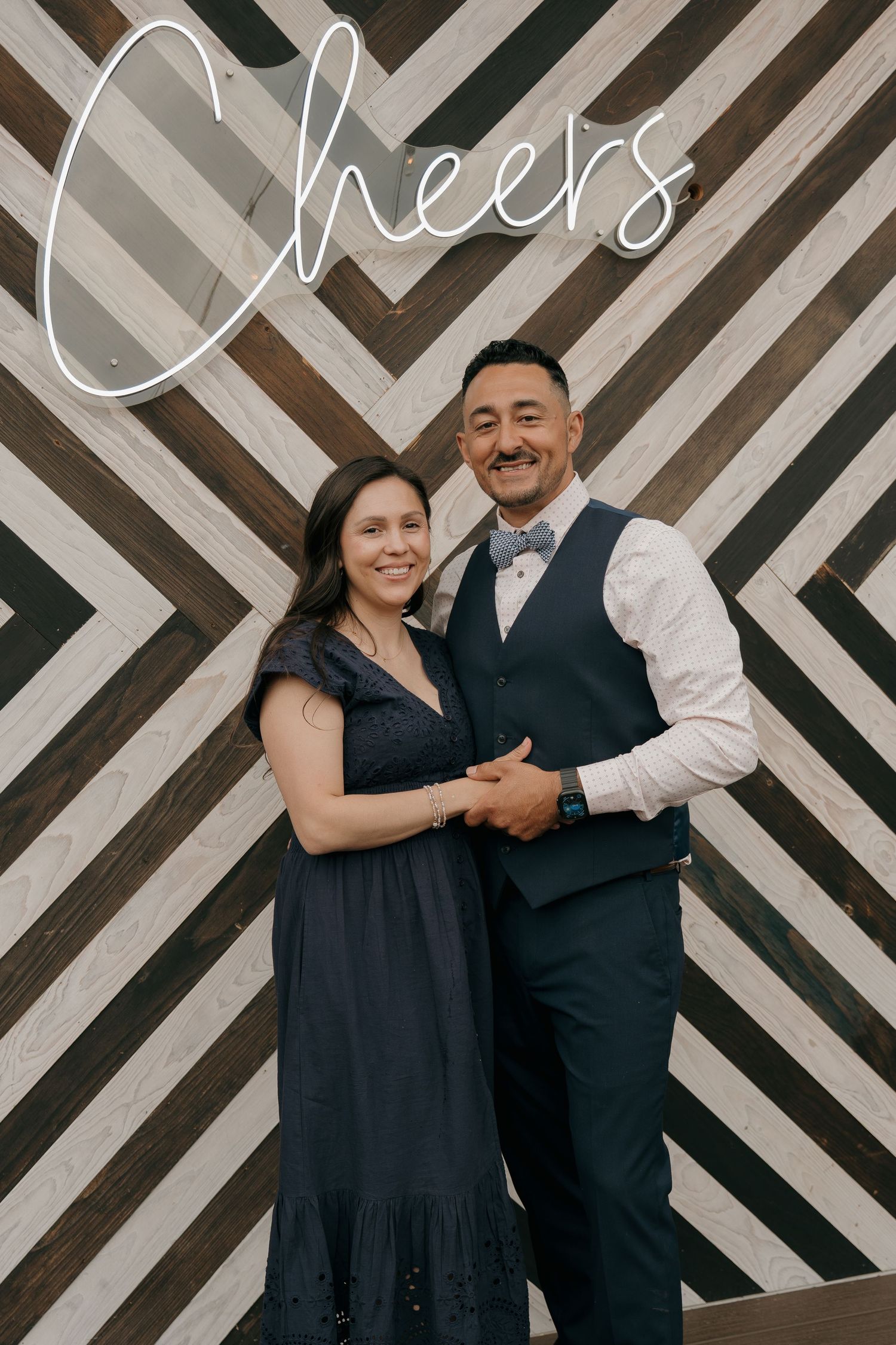 Couple posing together in front of striped chevron wood backdrop with glowing Cheers neon sign.