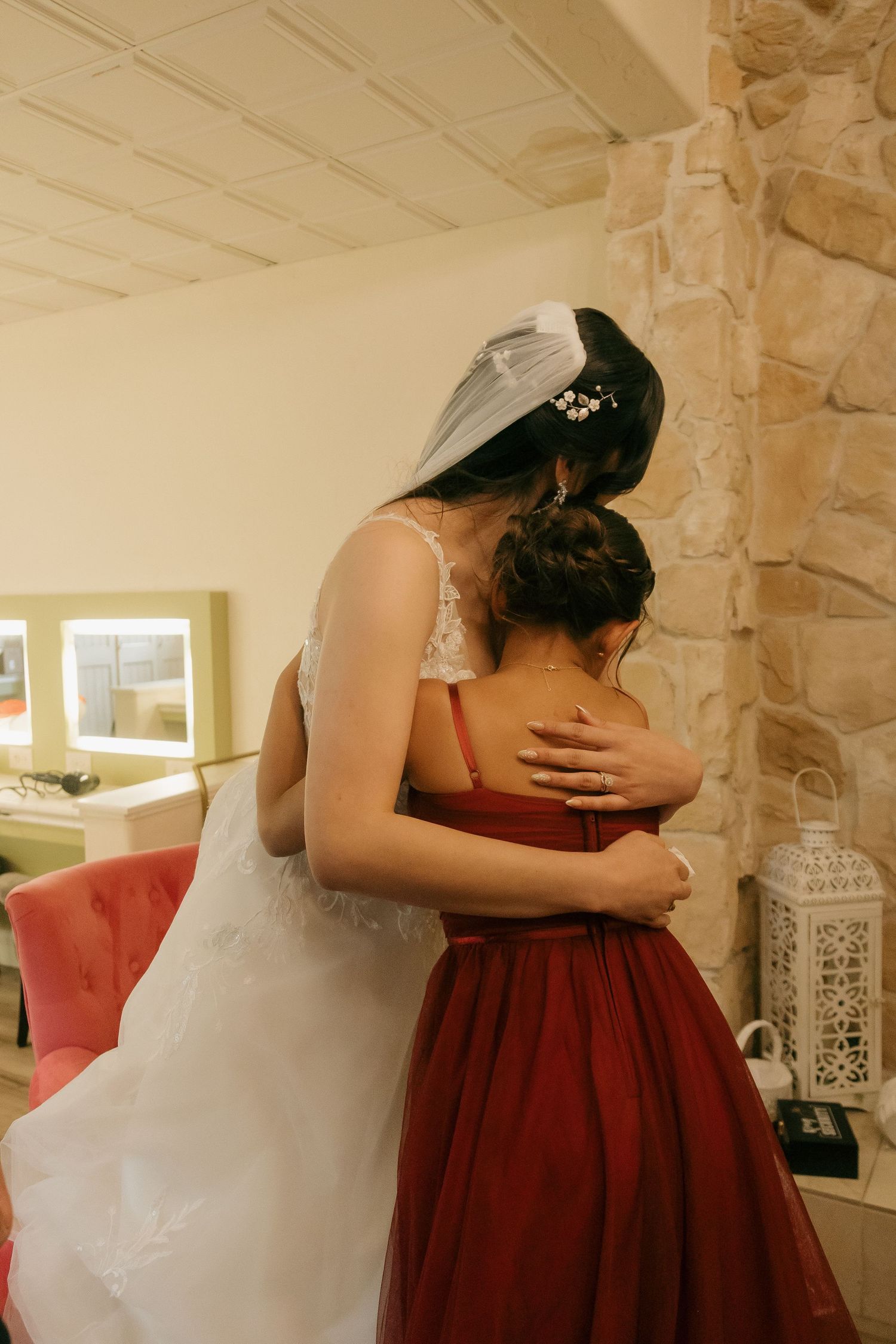 Bride in white gown shares a heartfelt hug with a young girl in a red dress indoors.