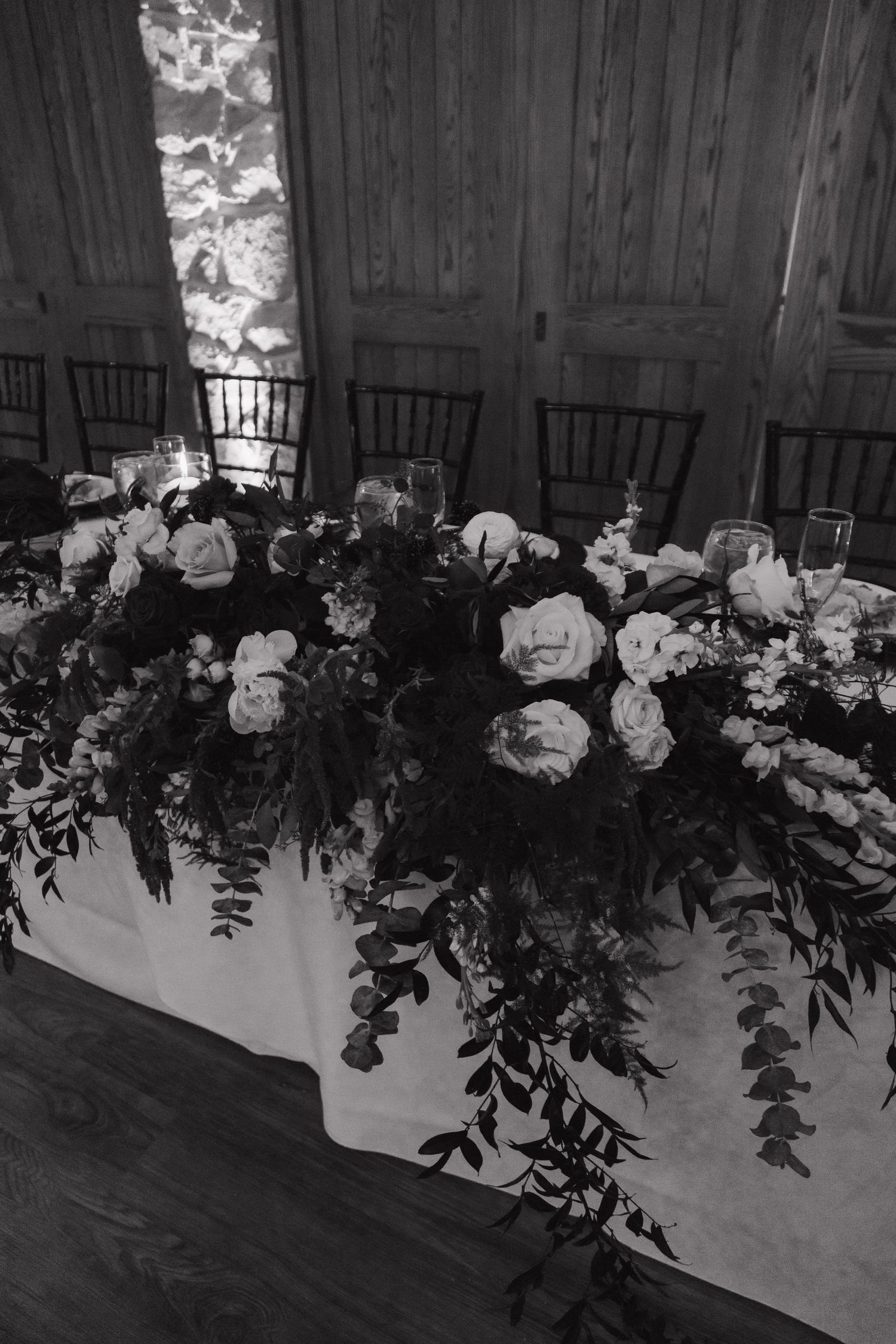 Black and white photo of lush floral centerpiece with roses and eucalyptus on white draped head table.