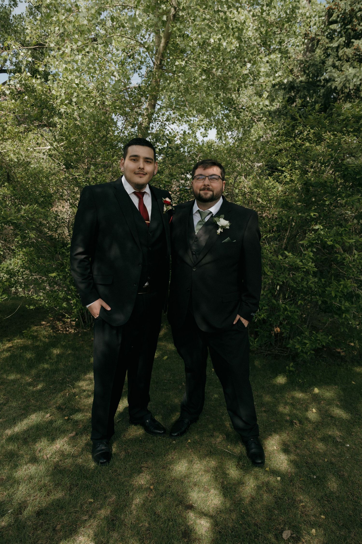 Two groomsmen in black suits with boutonnieres pose outdoors among green trees at a wedding.