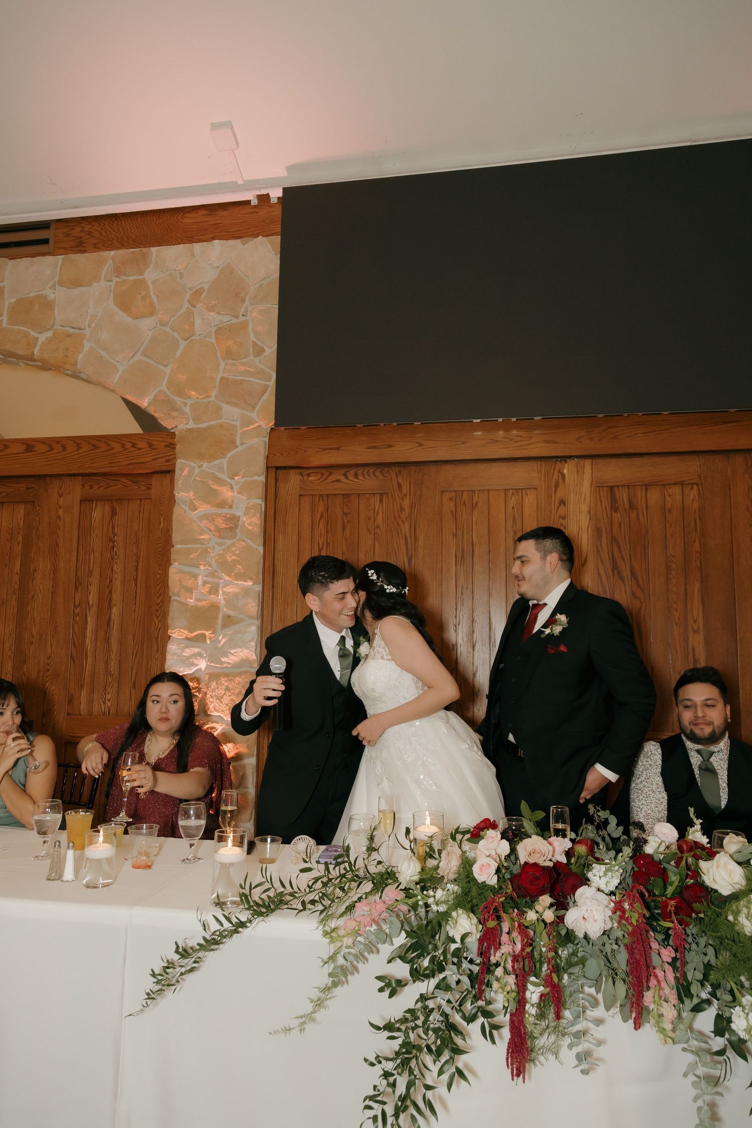 Groom gives speech at wedding reception head table adorned with red floral arrangements and candles.