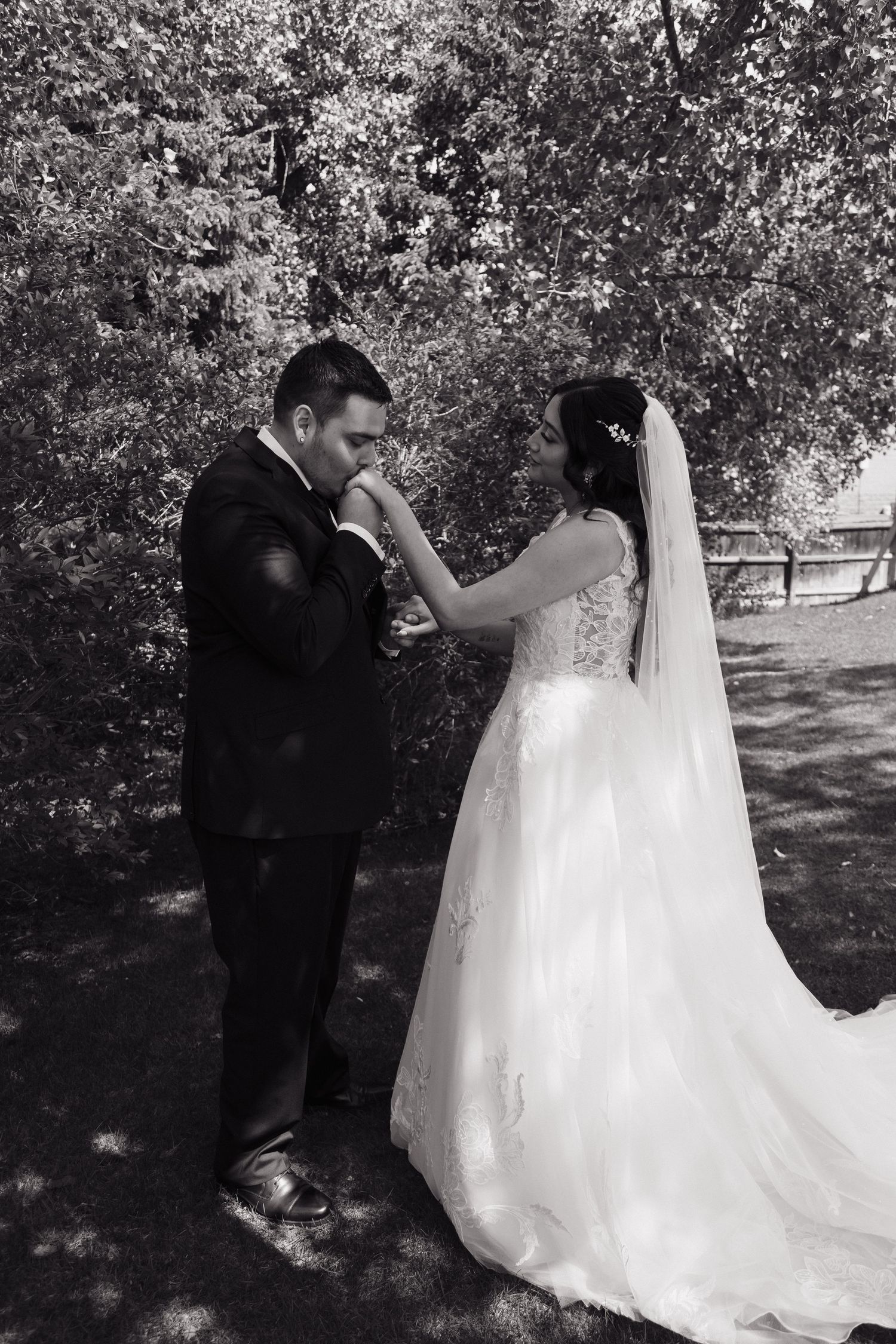 Groom kissing bride's hand in a romantic black and white wedding photo outdoors among lush trees.