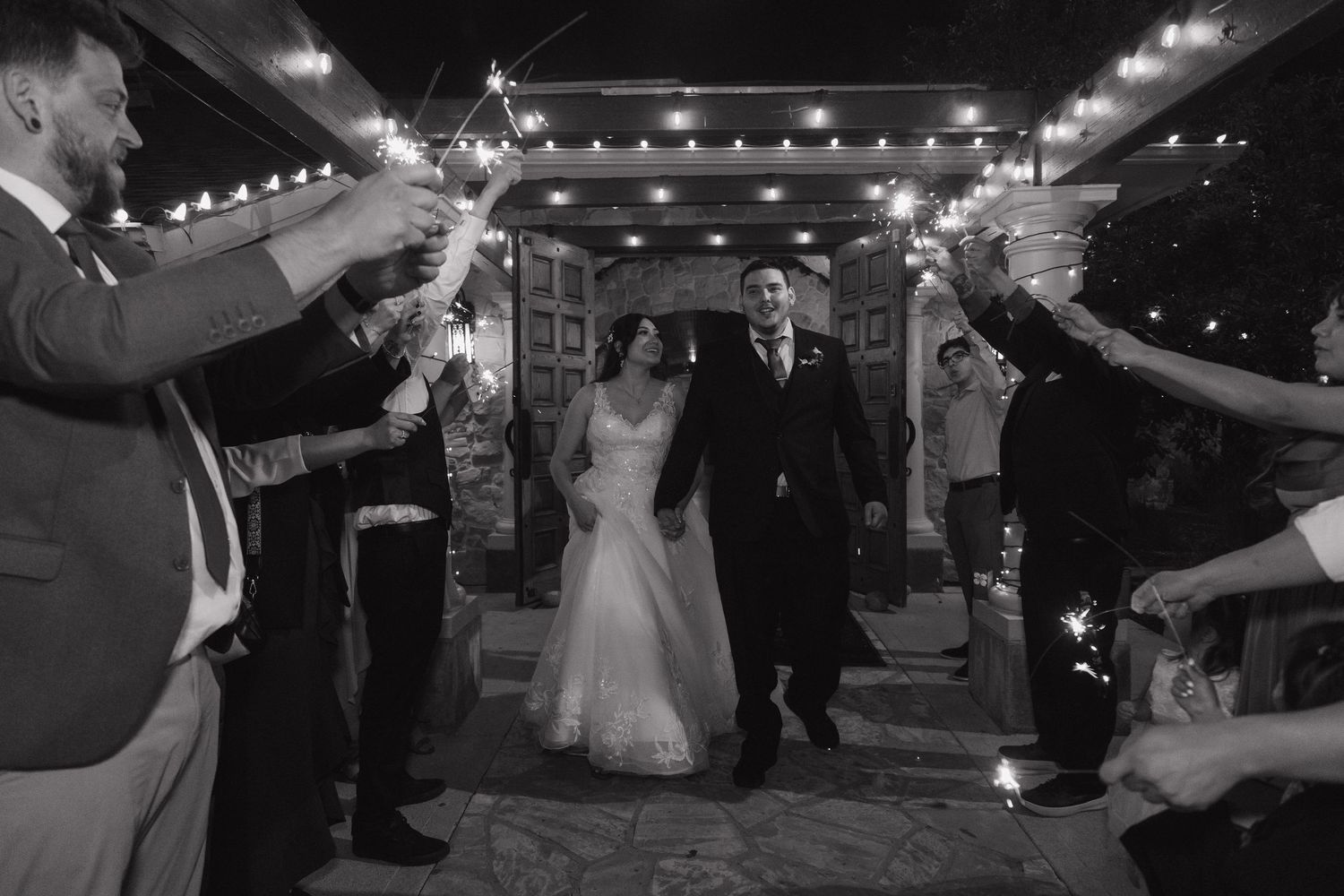 Black and white photo of bride and groom exiting wedding reception through sparkler tunnel with cheering guests.