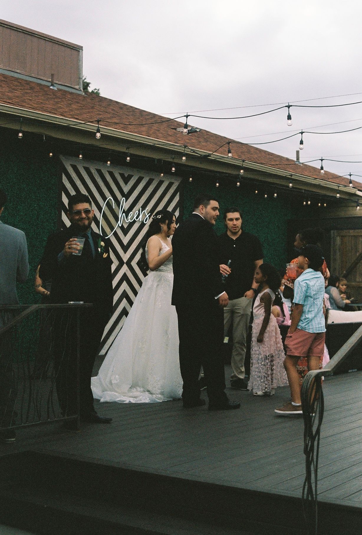 Bride and groom mingle with guests near Cheers backdrop at outdoor wedding reception.