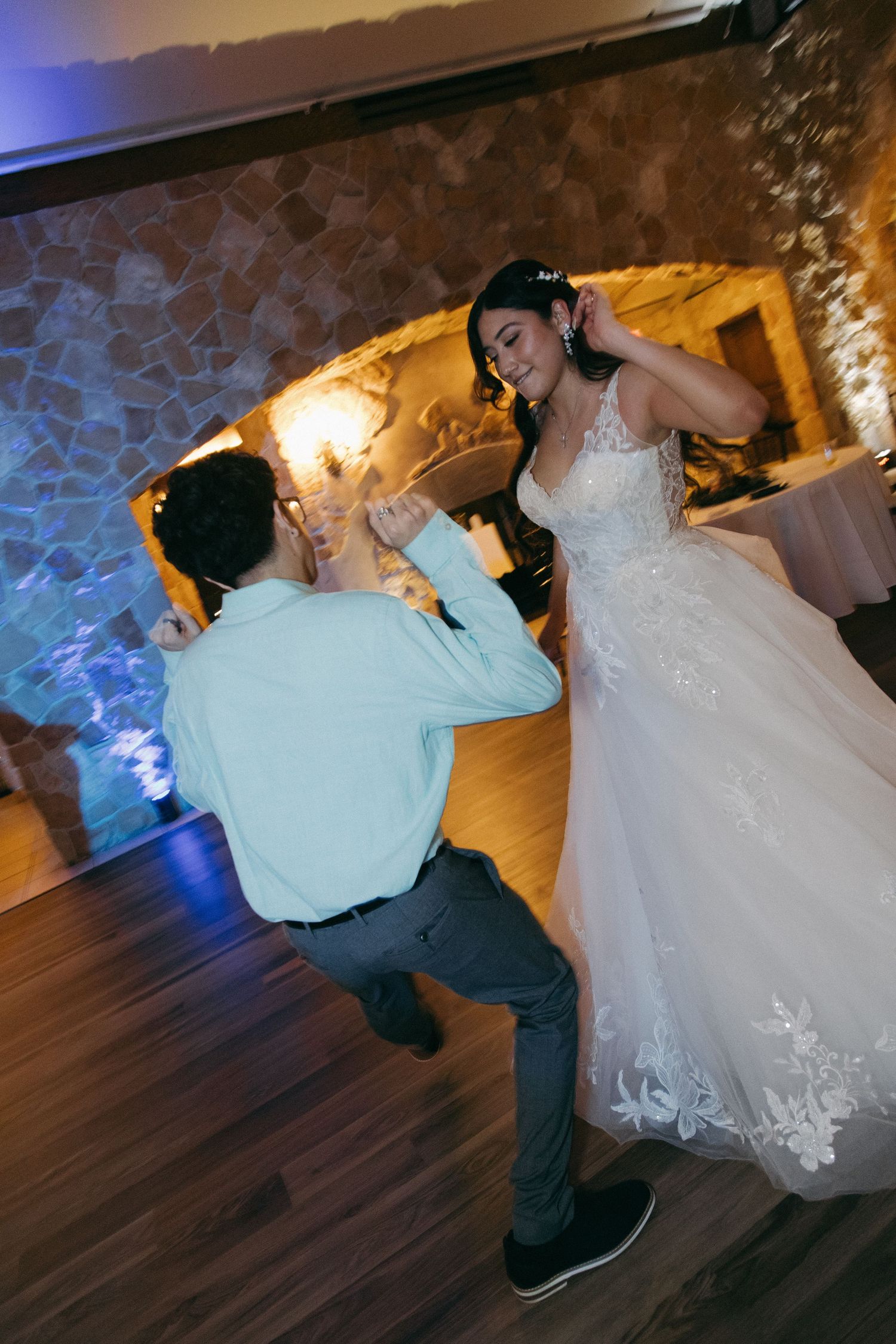 Bride in white ballgown dances with guest in mint shirt on dimly lit reception dance floor.