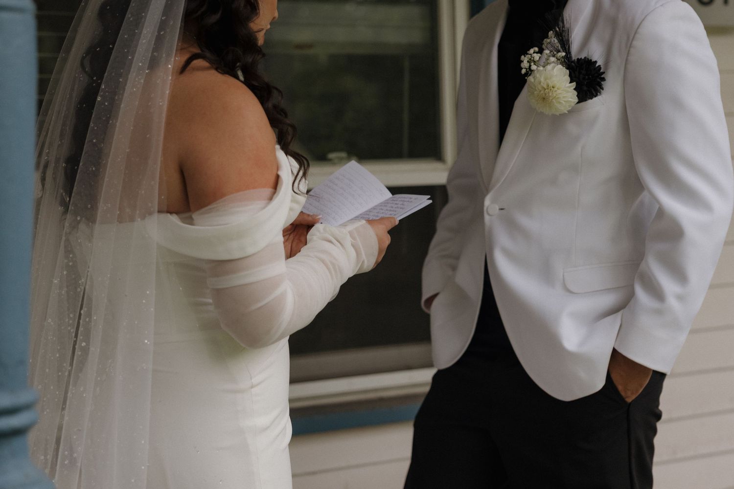 Bride in white strapless gown and veil reads wedding vows while groom in white suit with boutonniere listens.