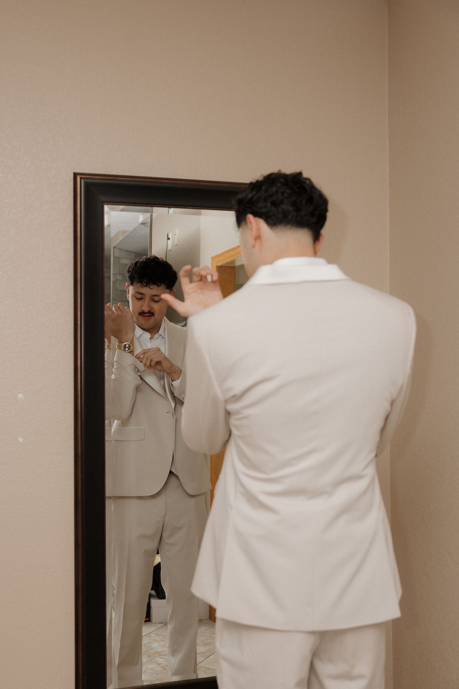A groom in a white suit adjusts his tie while looking at his reflection in a mirror before his wedding.