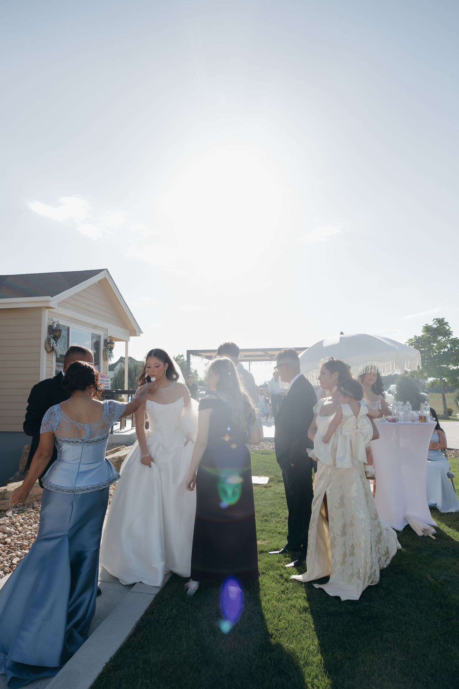 Wedding party and guests gathered outdoors in bright sunlight, bride in white gown surrounded by attendants.