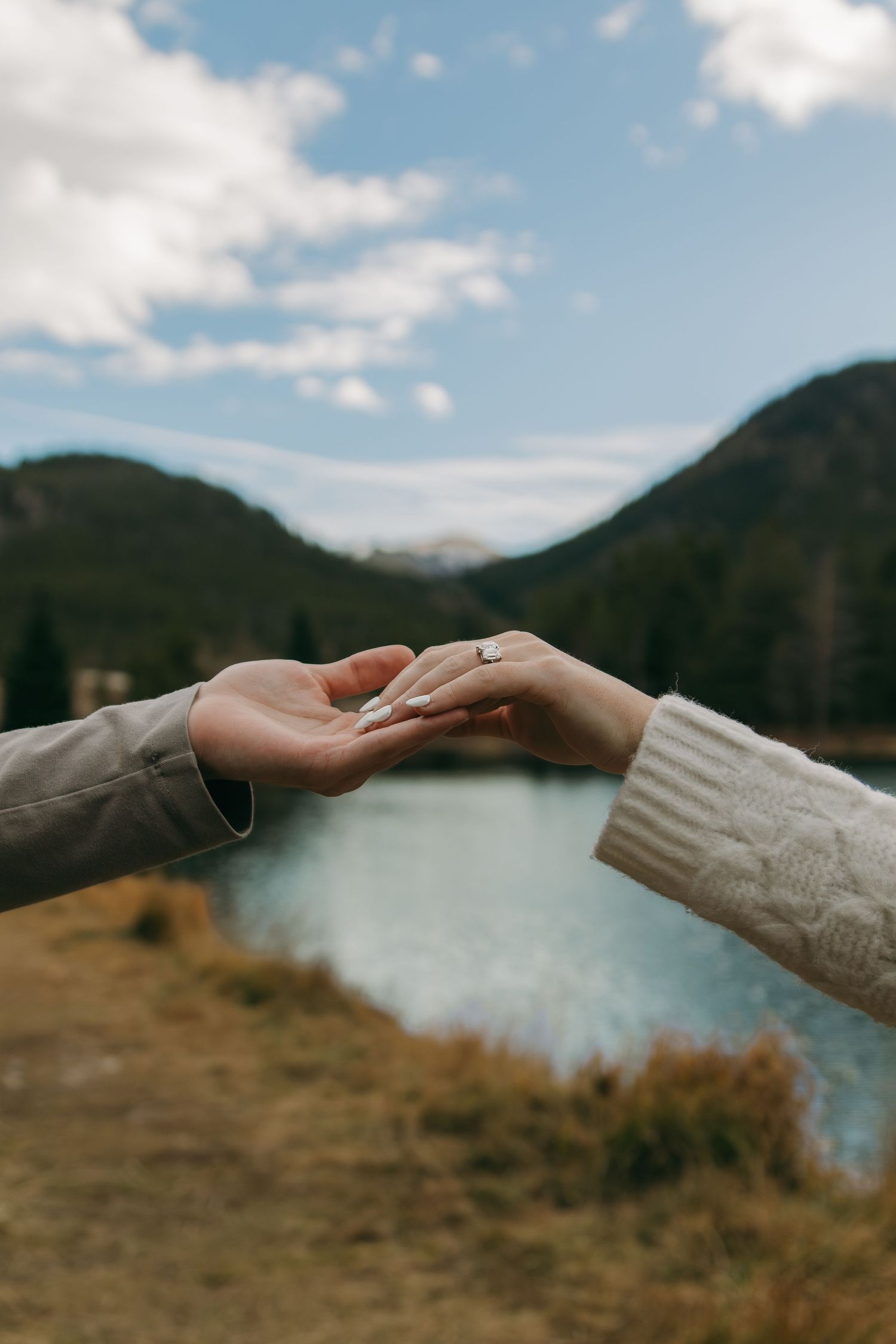 Two hands reach across a scenic mountain lake landscape with blue skies and rugged peaks in the background.
