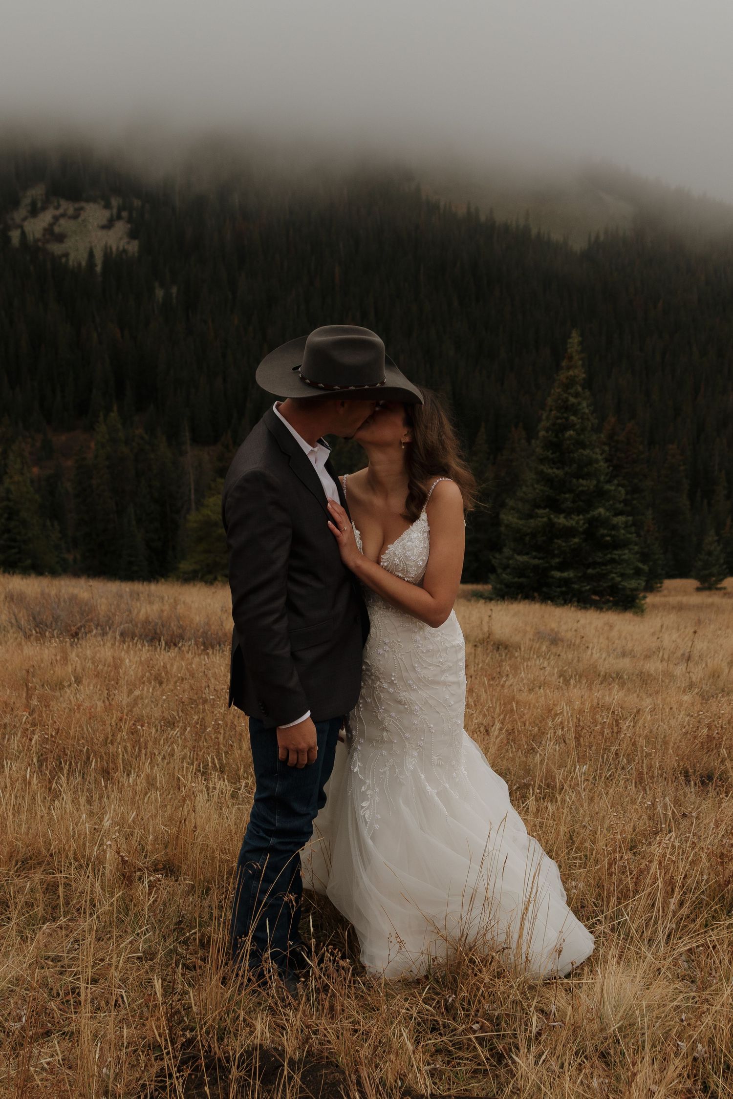 A romantic wedding couple embraces in a misty mountain meadow surrounded by evergreen trees.