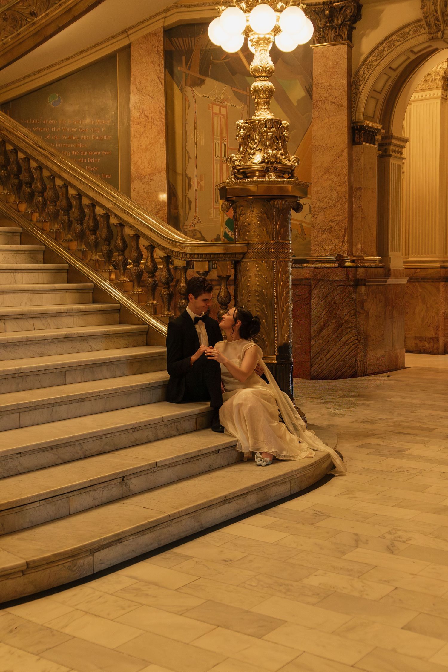Wedding couple sitting on marble steps in an ornate historic building interior.