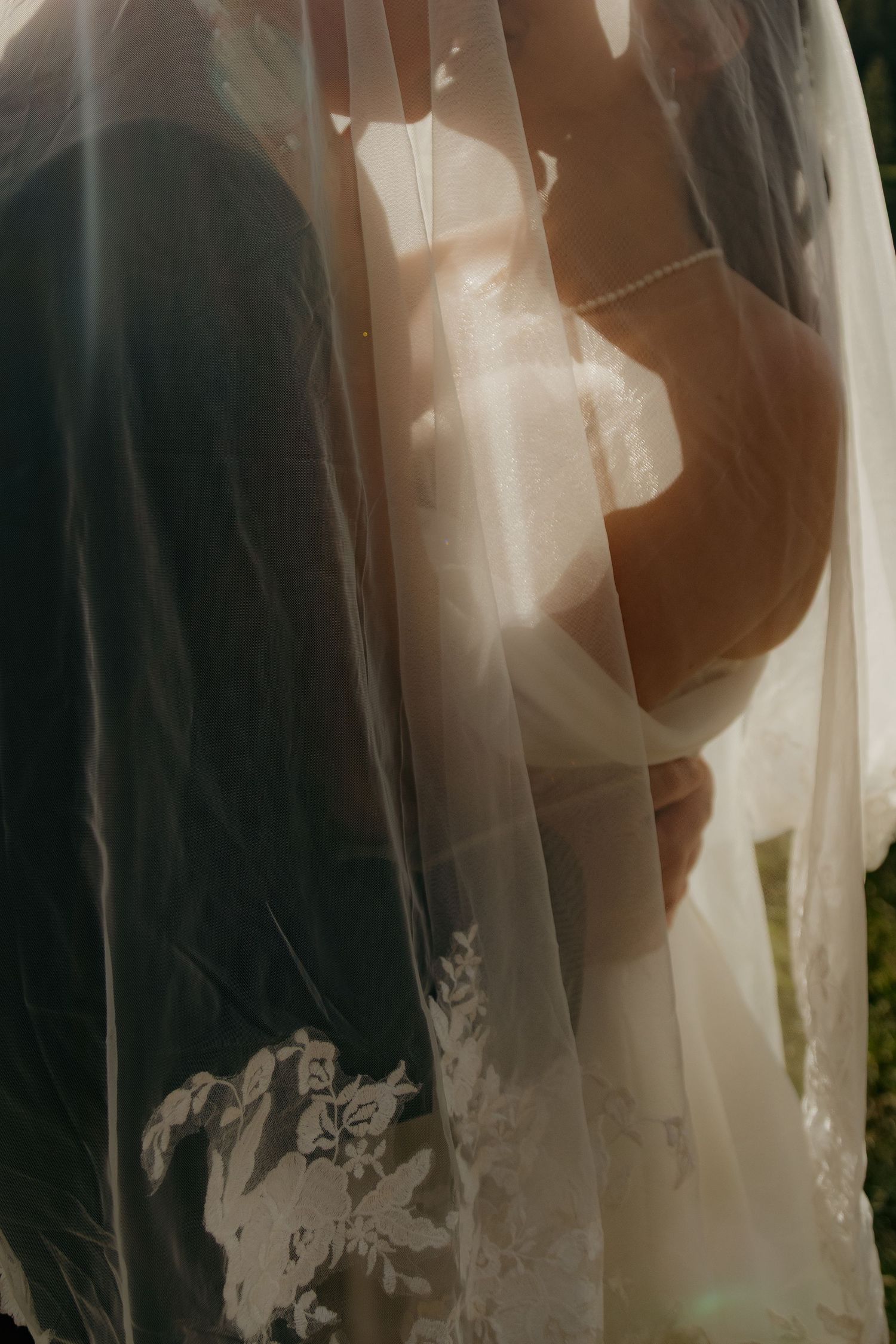 A bride and groom share an intimate moment under a lace wedding veil, backlit by warm golden sunlight.