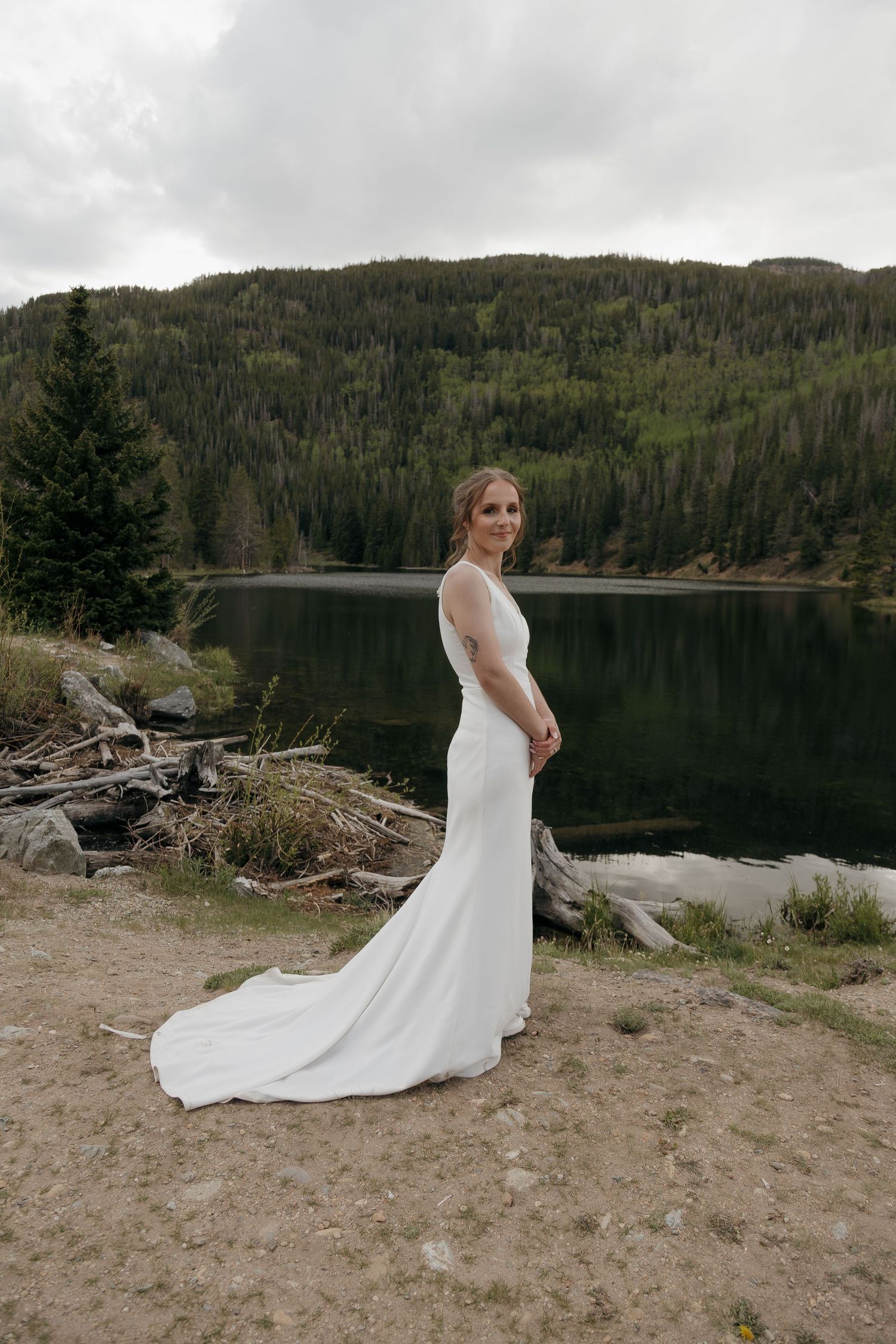 Bride in elegant white gown with train poses beside serene mountain lake on overcast day.