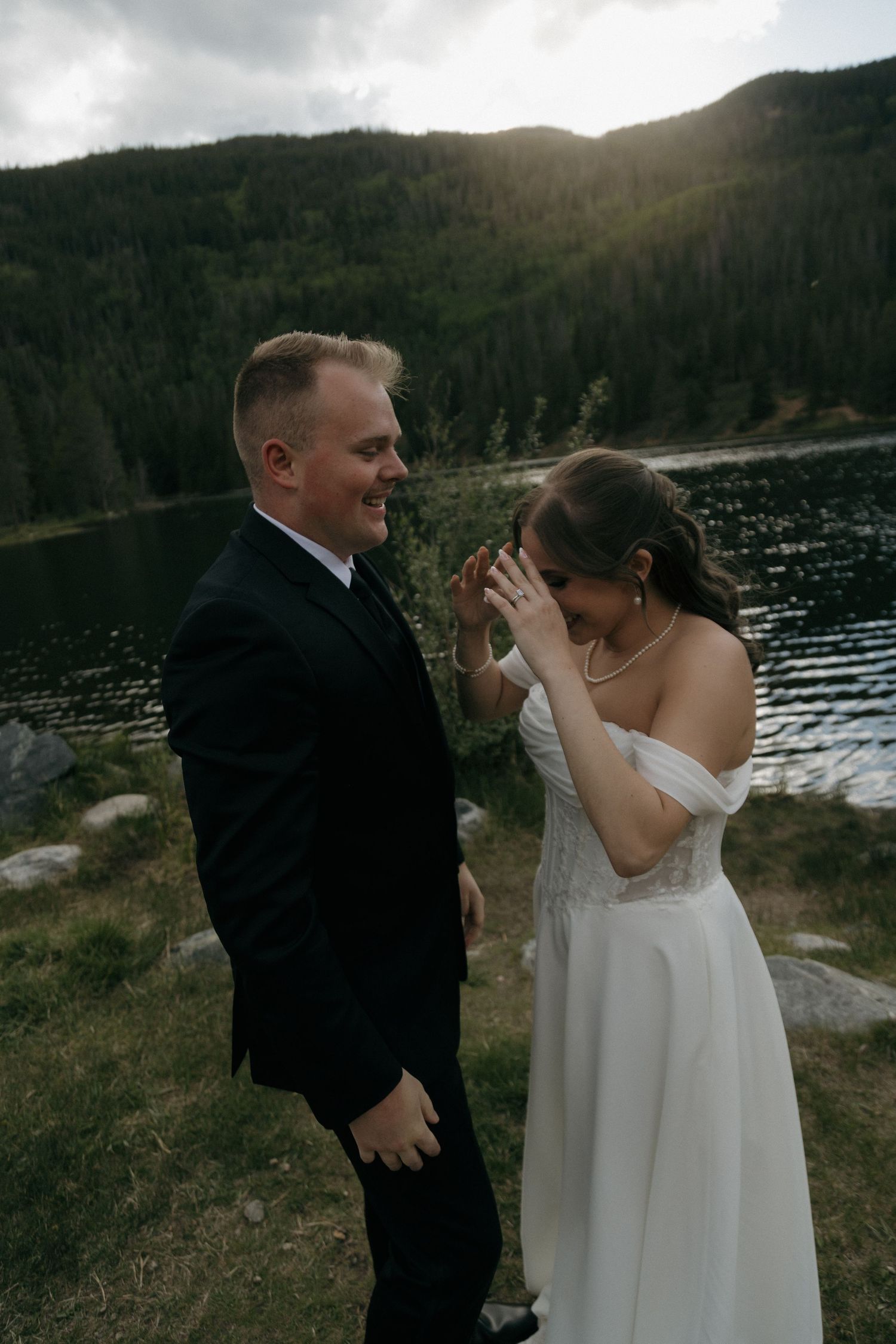 Groom smiling as emotional bride covers face with ringed hand near scenic mountain lake.