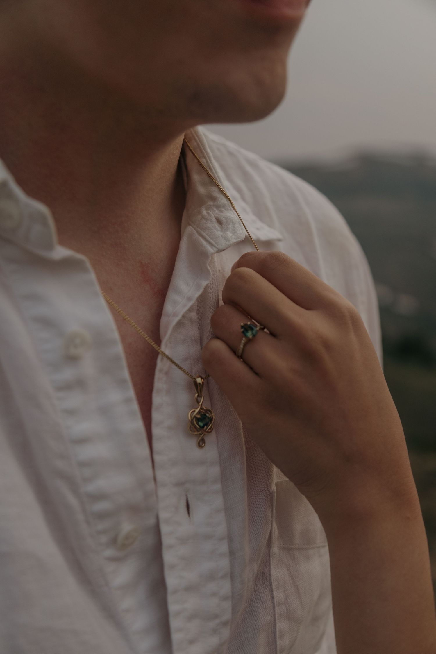 Man in white linen shirt holding necklace pendant, wearing turquoise gemstone ring, with scenic hills in background.