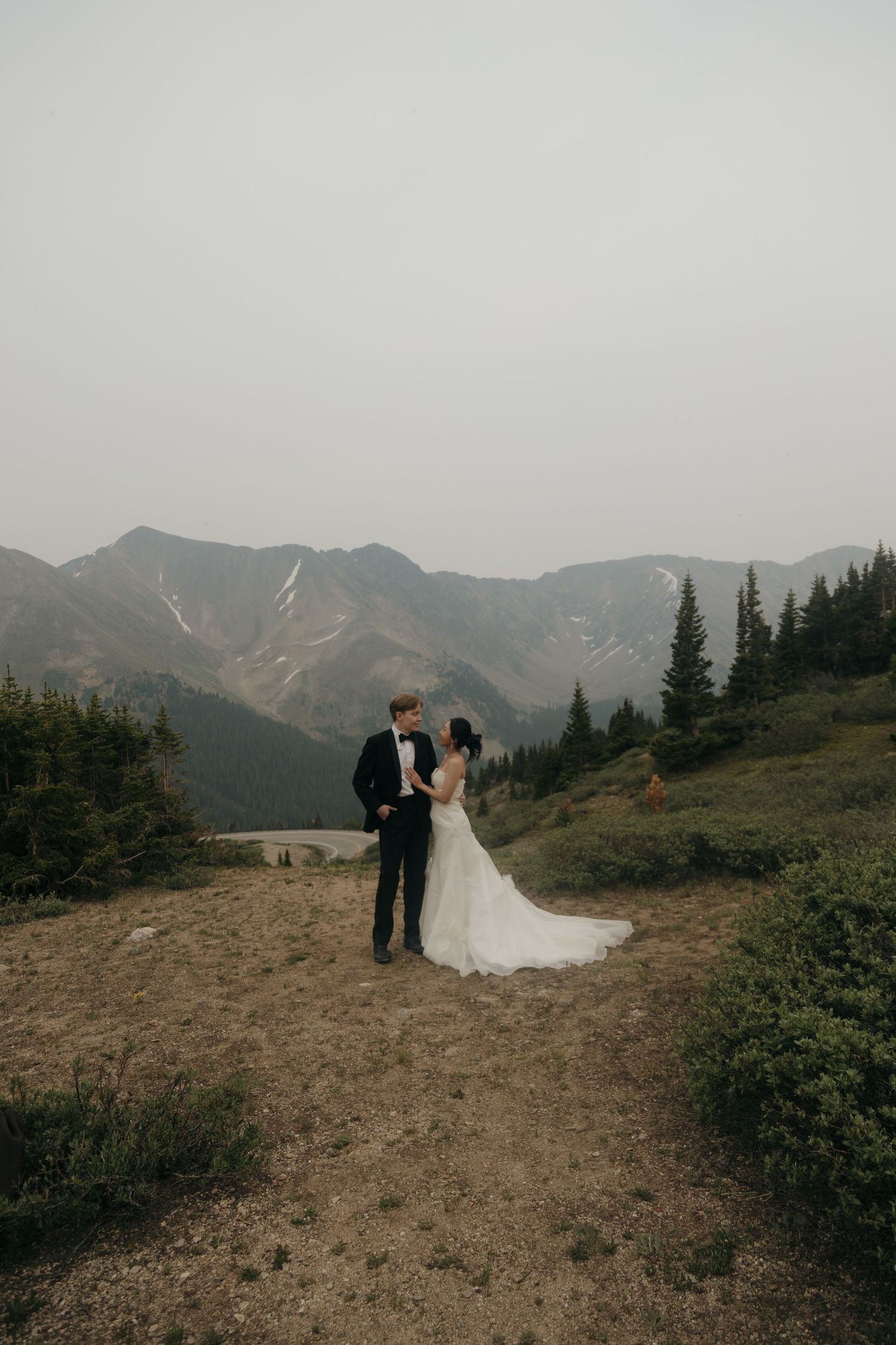 Wedding couple in formal attire stand together on a rocky mountain path with misty peaks behind them.