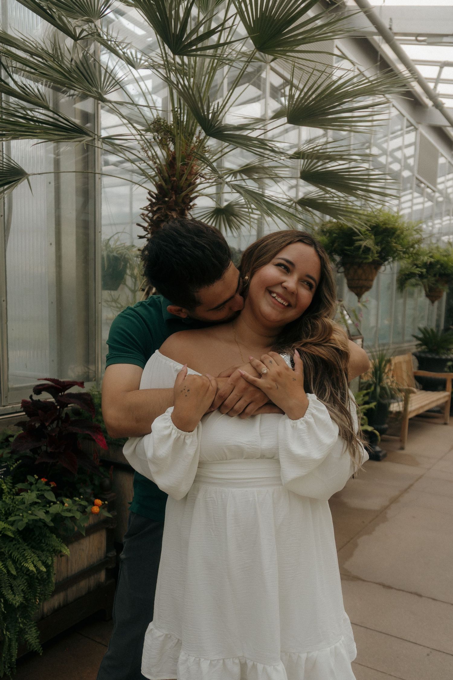 Couple embracing in a lush greenhouse, man hugging woman in white dress from behind as she smiles joyfully.