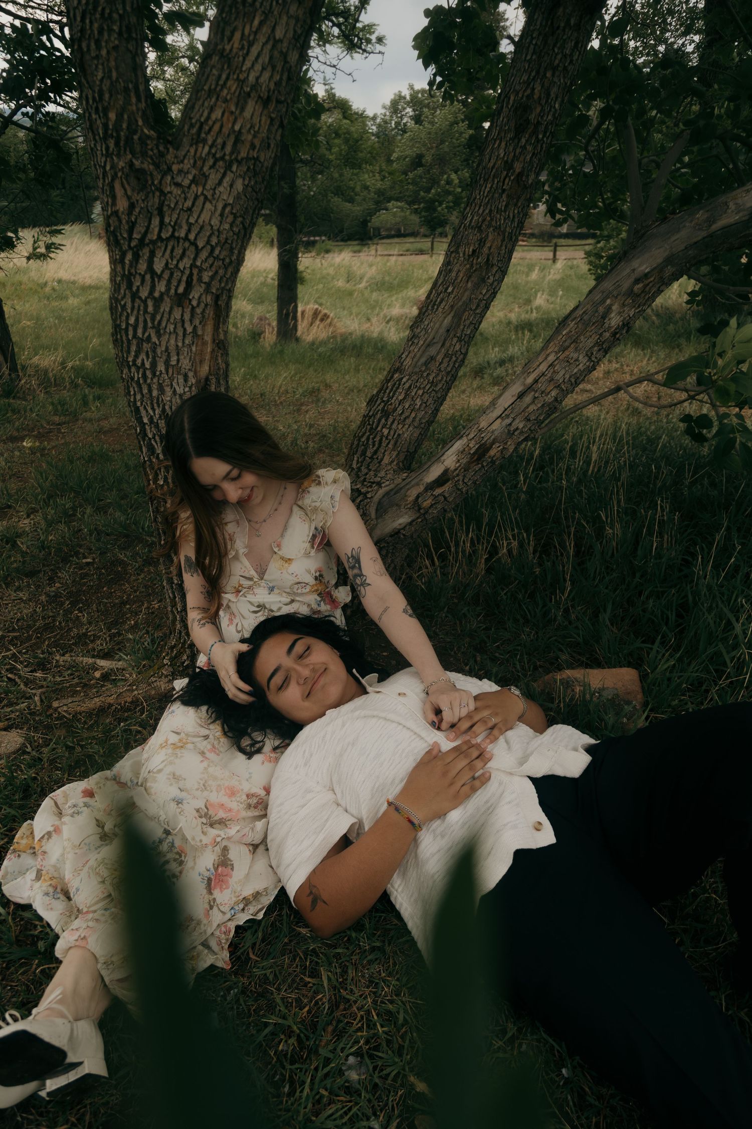 Couple relaxing under trees in a field, woman in floral dress cradling partner lying in her lap.