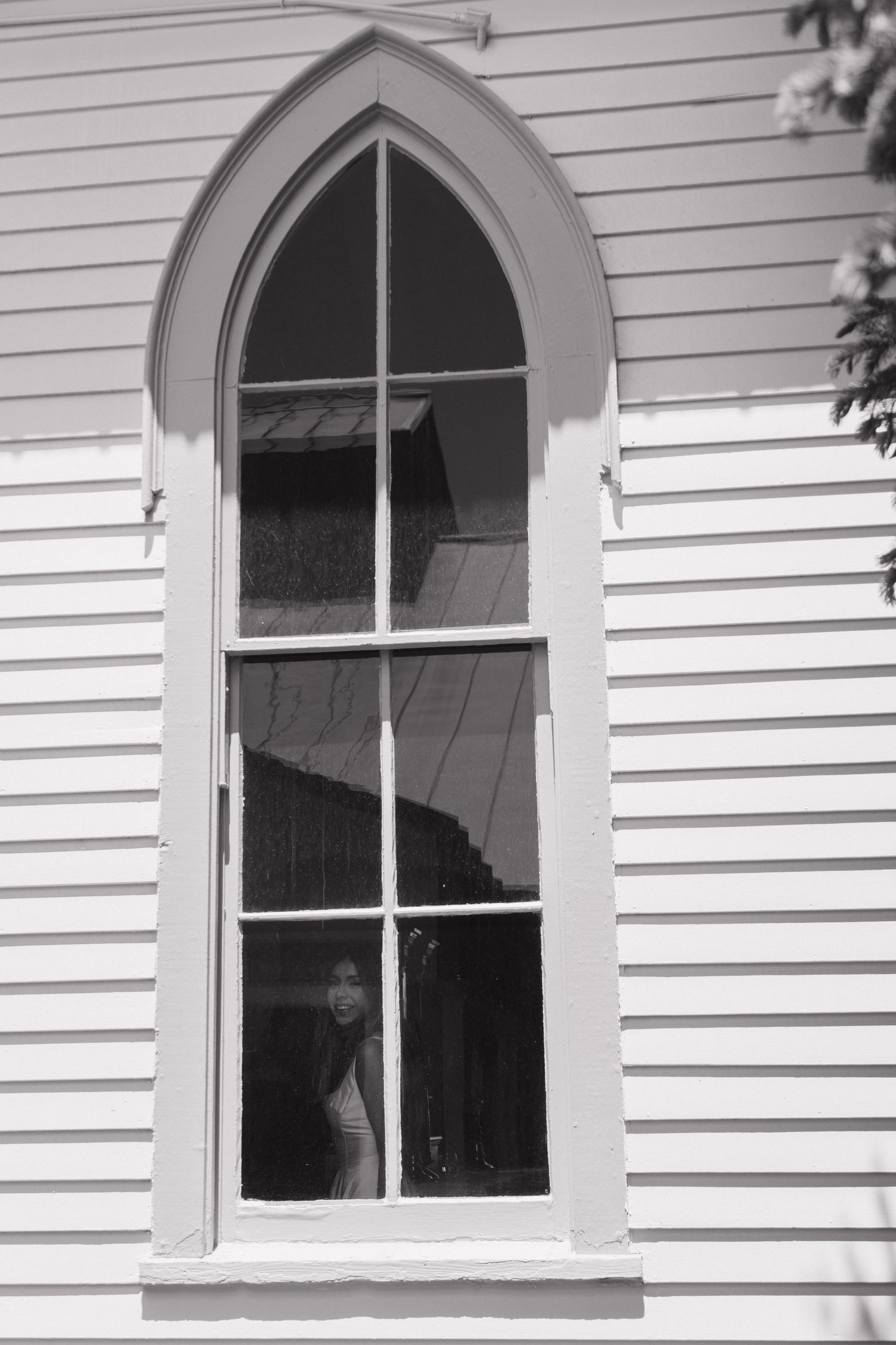 Black and white photo of a bride peering through a Gothic arched church window.