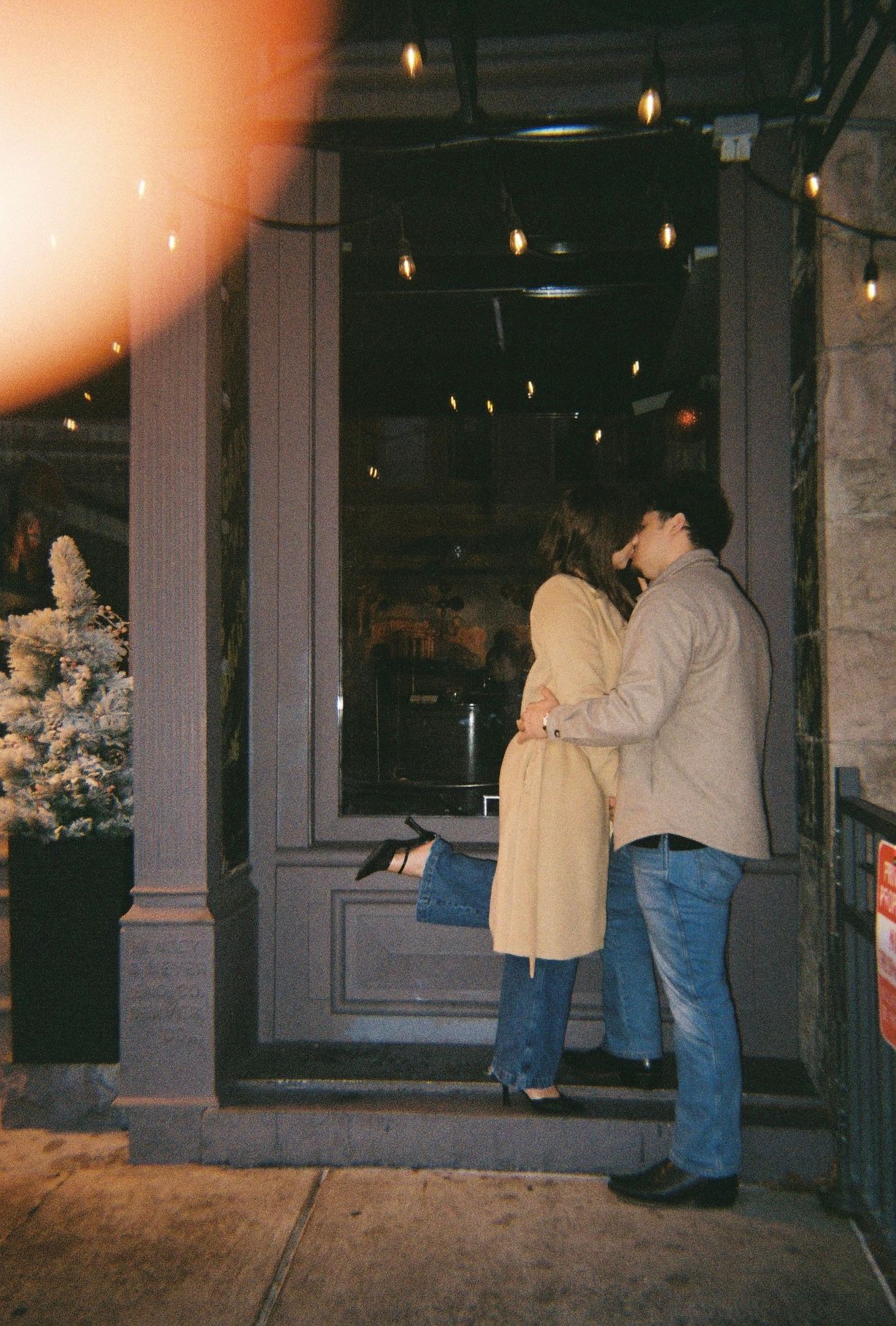 A romantic couple dances together on a dimly lit sidewalk outside a building with glowing lights in the background.