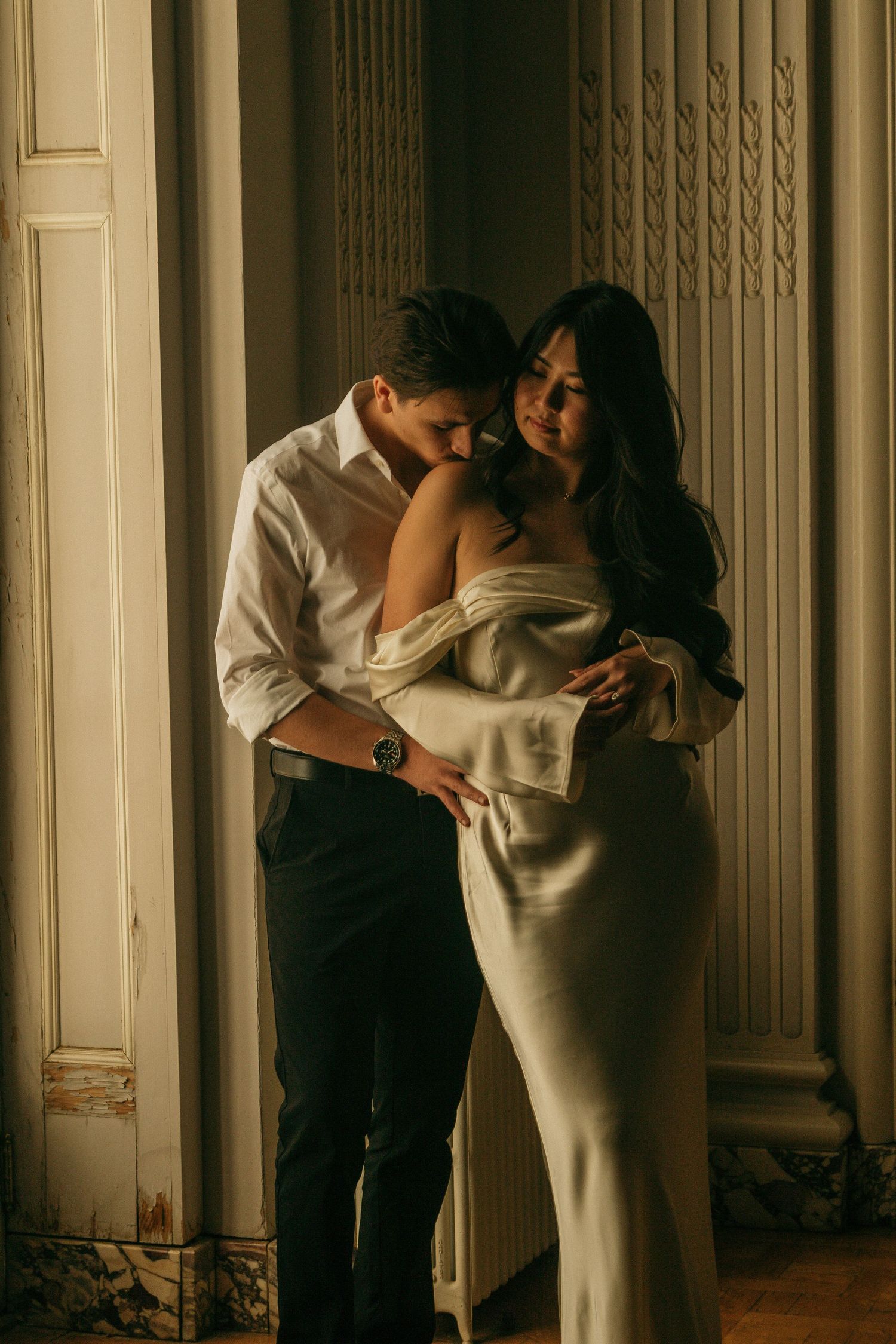 Romantic couple embraces in elegant white dress and formal attire against ornate interior wall.