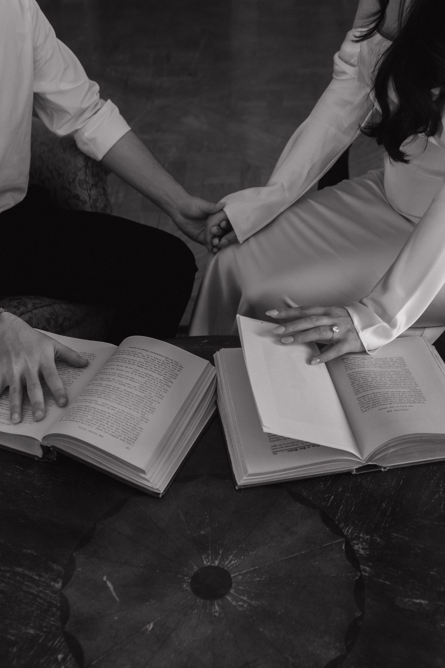 Hands turning pages of an open book on a dark wooden surface in black and white.