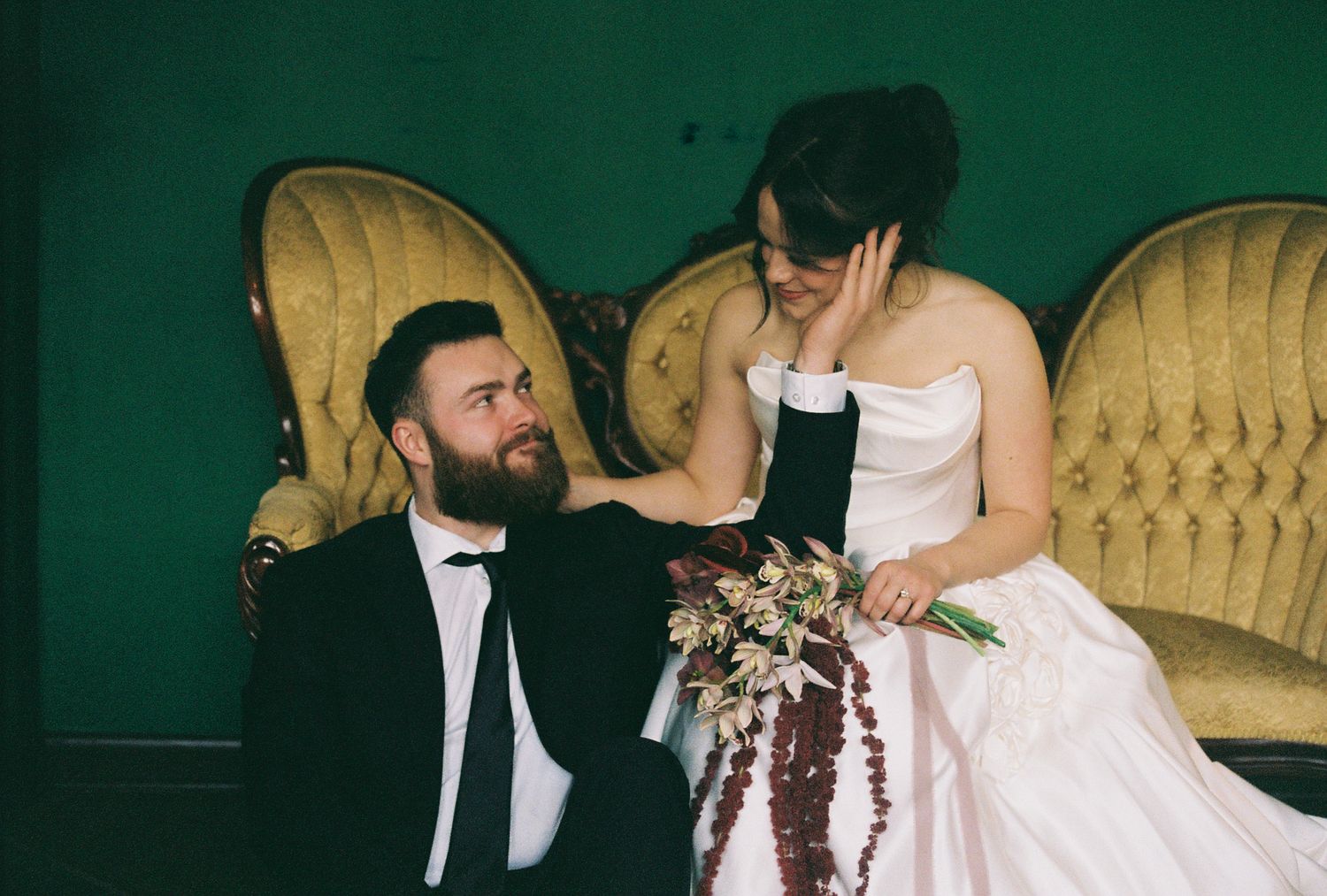 Wedding couple relaxing on vintage velvet couch against deep green backdrop.