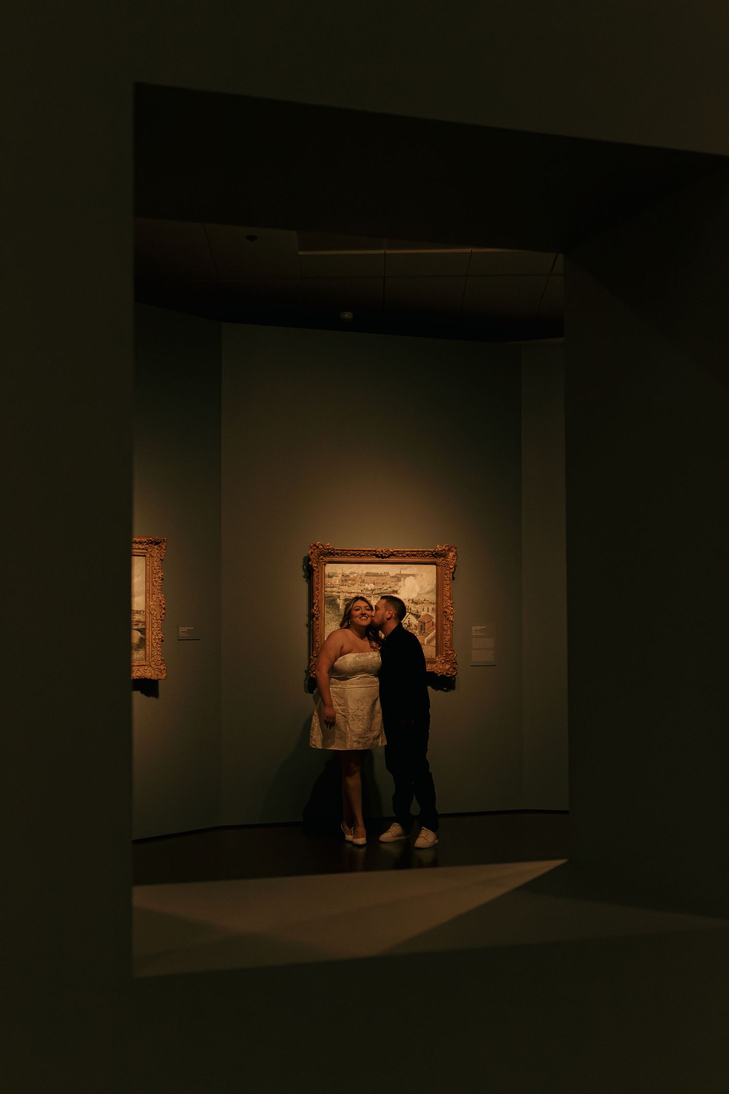 Museum visitors observe artwork in a dimly lit gallery space with spotlit paintings on dark walls.