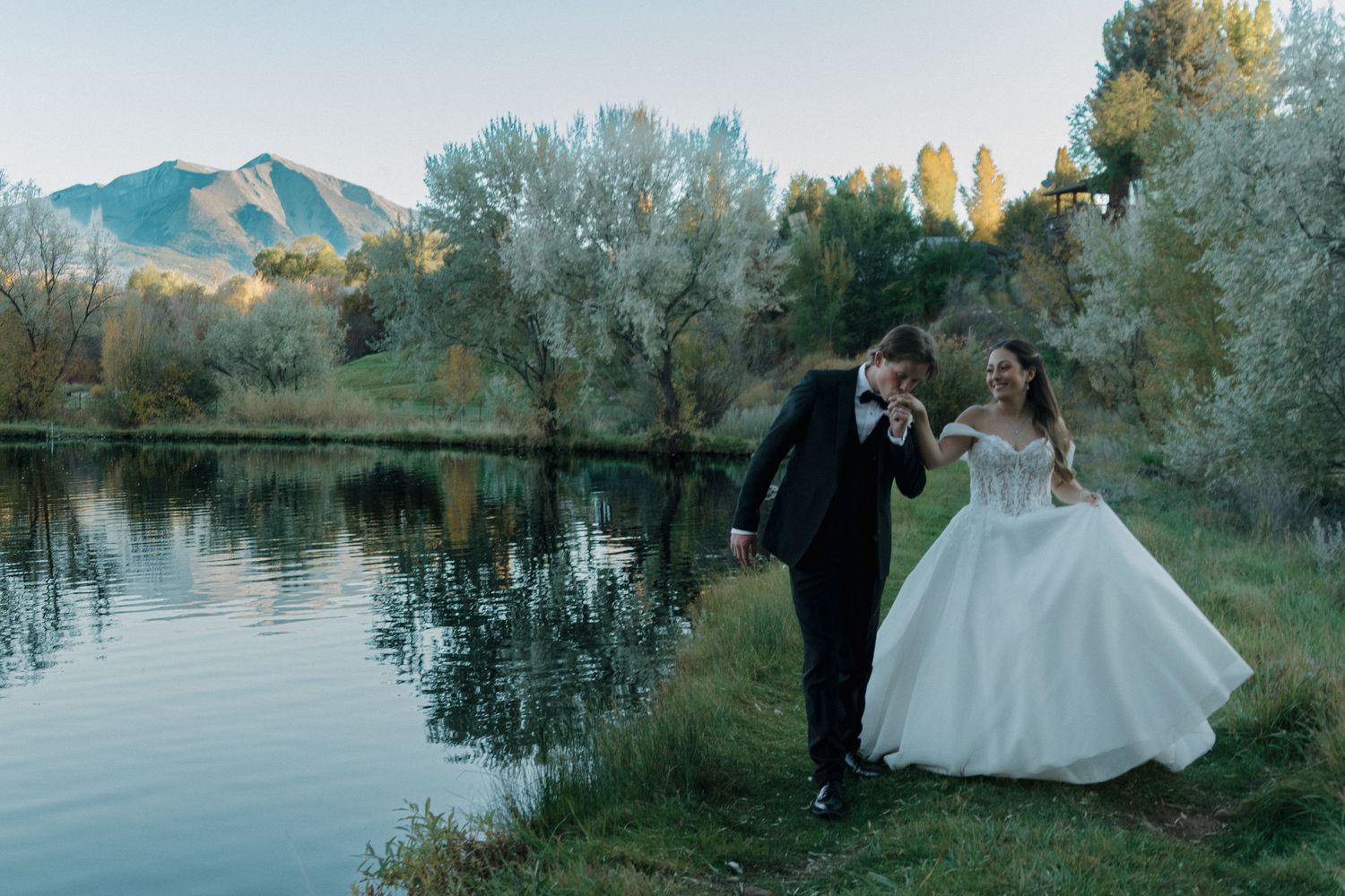 Wedding party poses by a serene lake at sunset with mountains in the background.