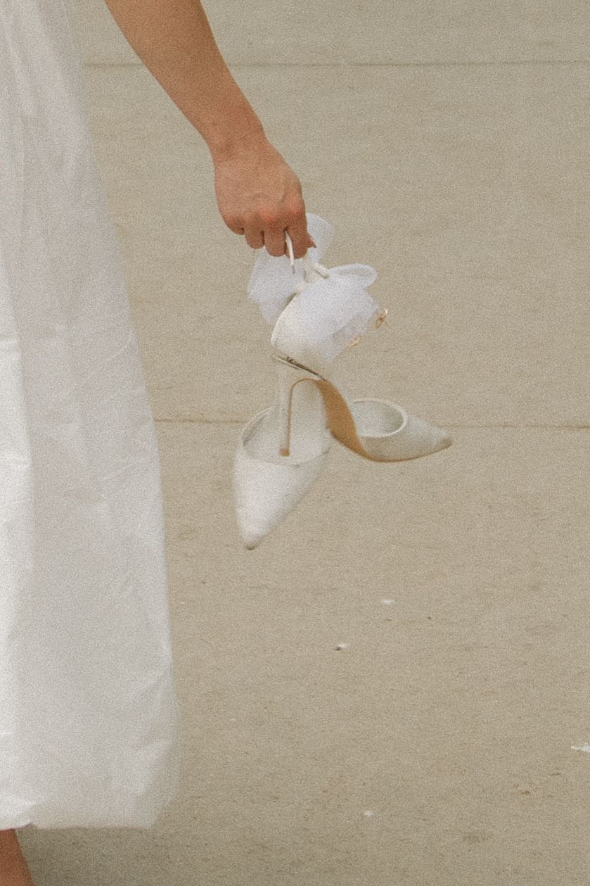 Person in white dress holding elegant white shoes against a light background.