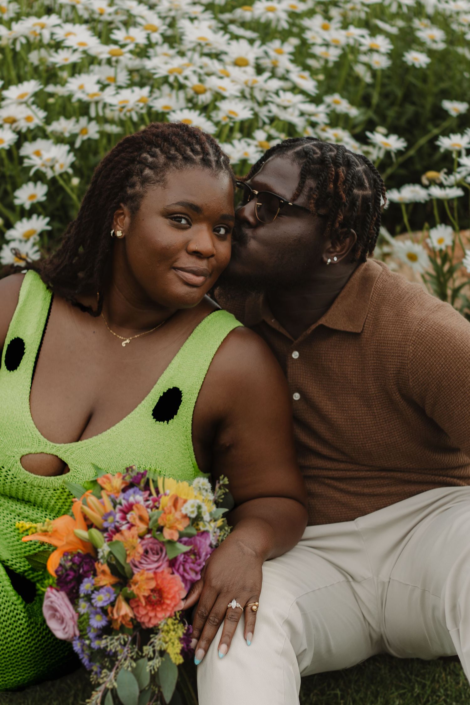 A couple sits in a field of daisies as they embrace while holding a vibrant bouquet of colorful flowers.