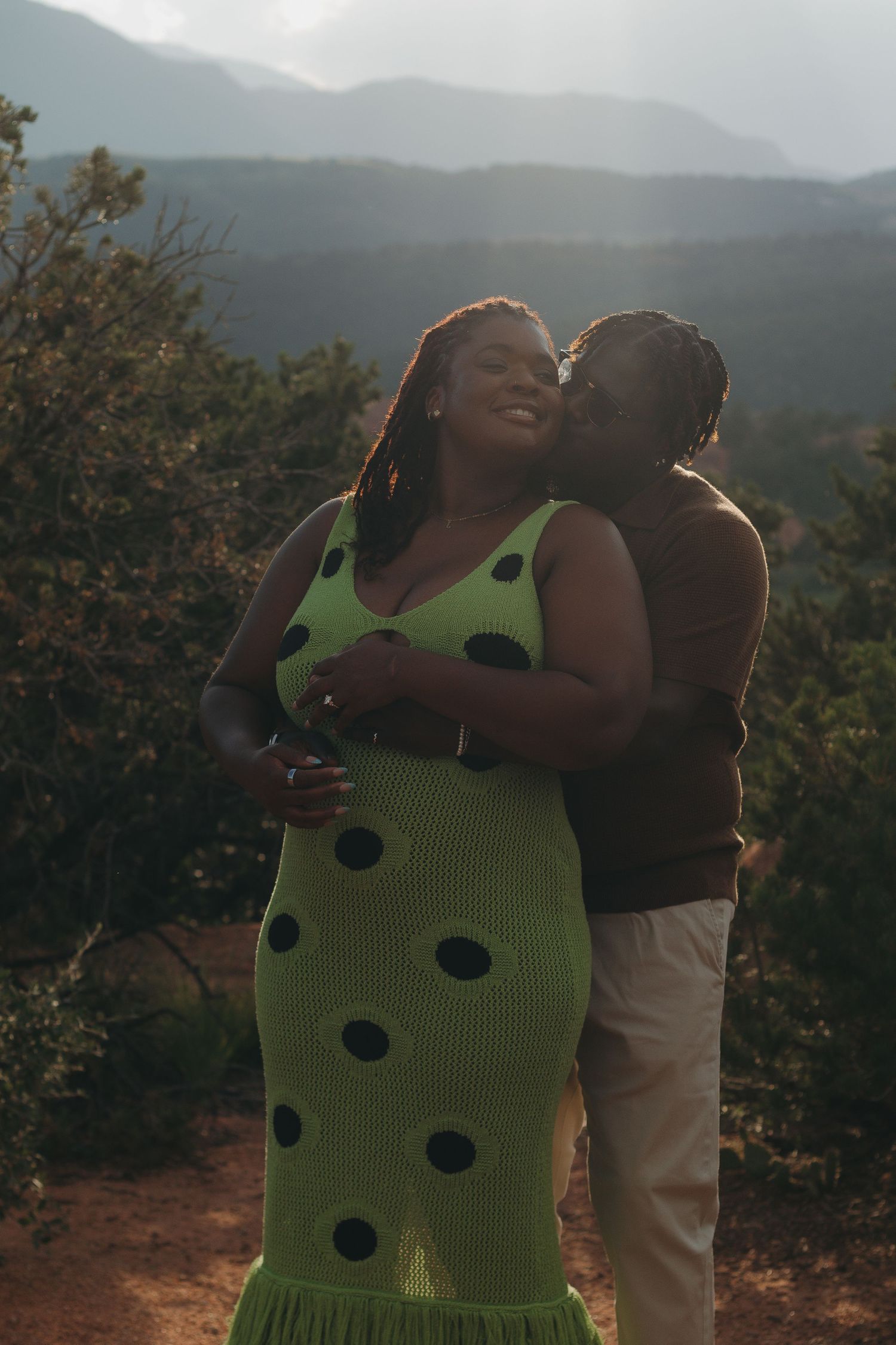 Couple embraces against a mountainous backdrop in warm evening light.