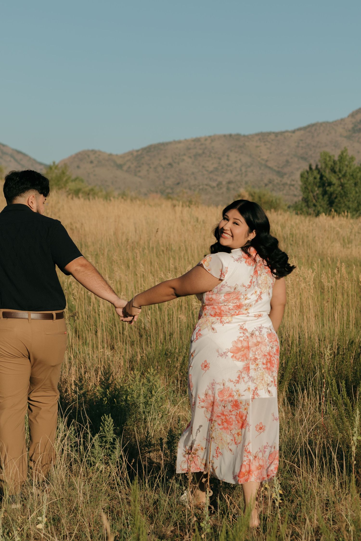 Couple walking hand in hand through tall grass with mountains in the background.